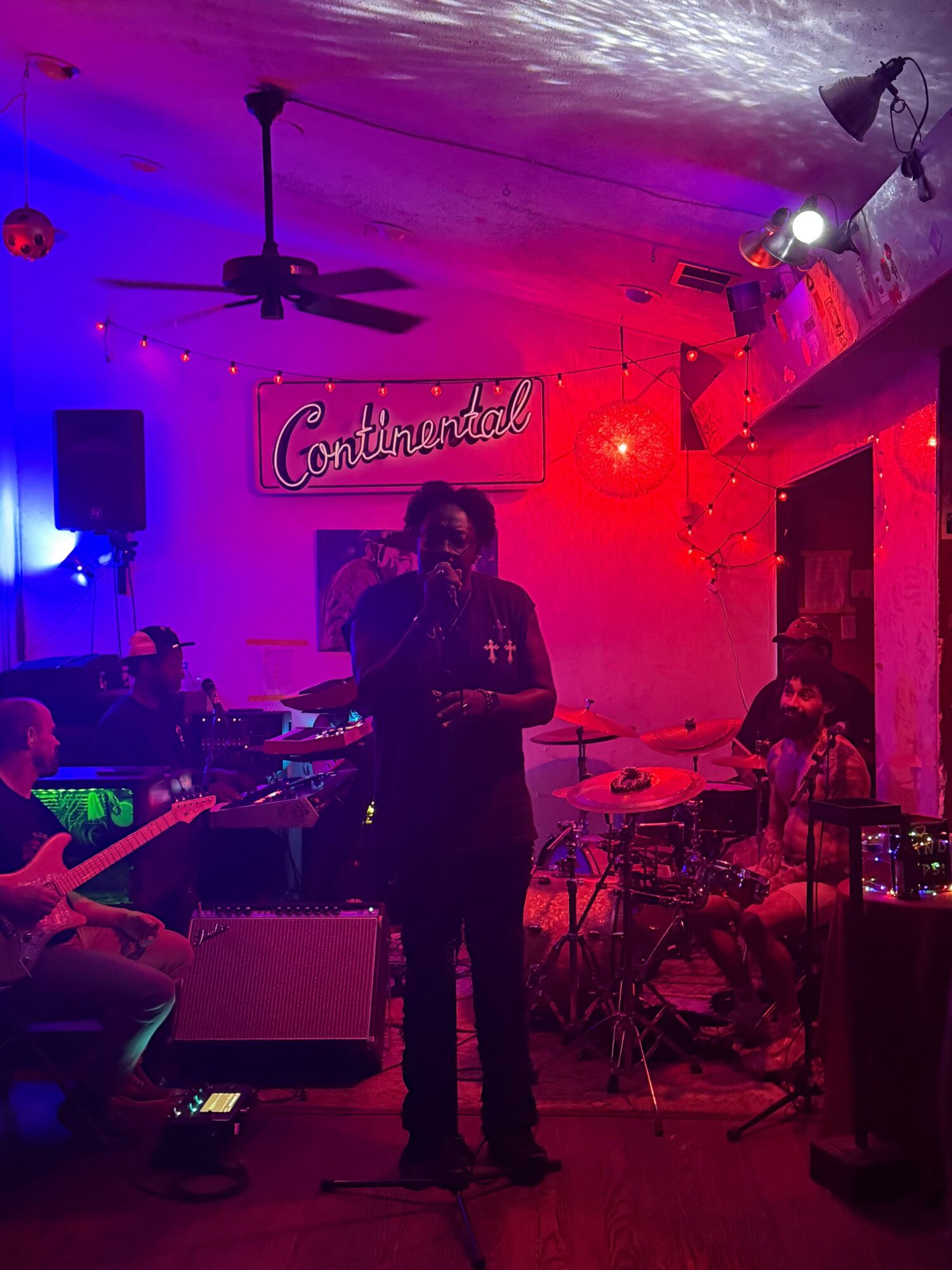 Person performing on stage with colorful lighting, musical instruments, and a sign reading 'Cactus' in the background.