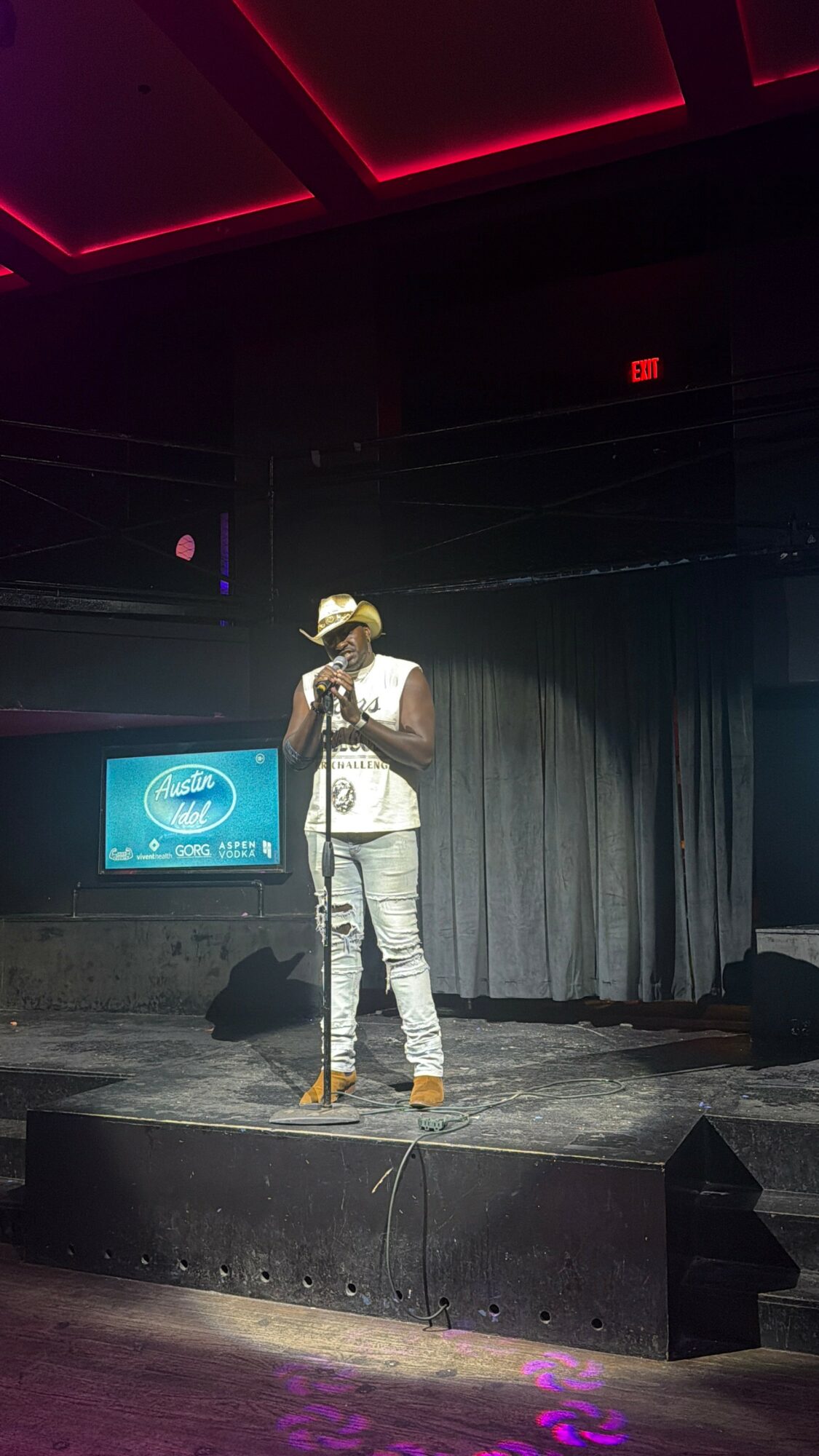 Person standing on stage with microphone, wearing a hat and casual clothing, in front of a curtain and screen.