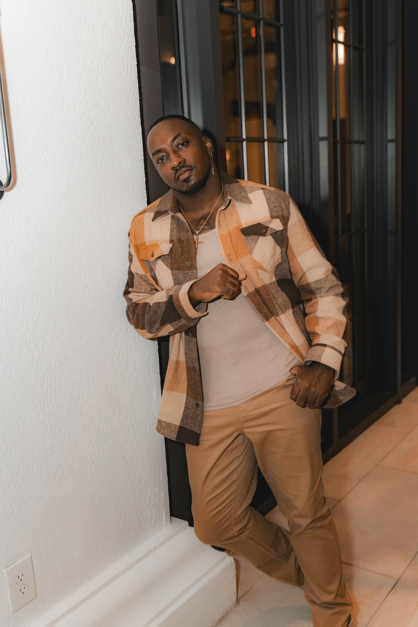 Man leaning against a wall in a modern interior, wearing a plaid shirt and beige pants.