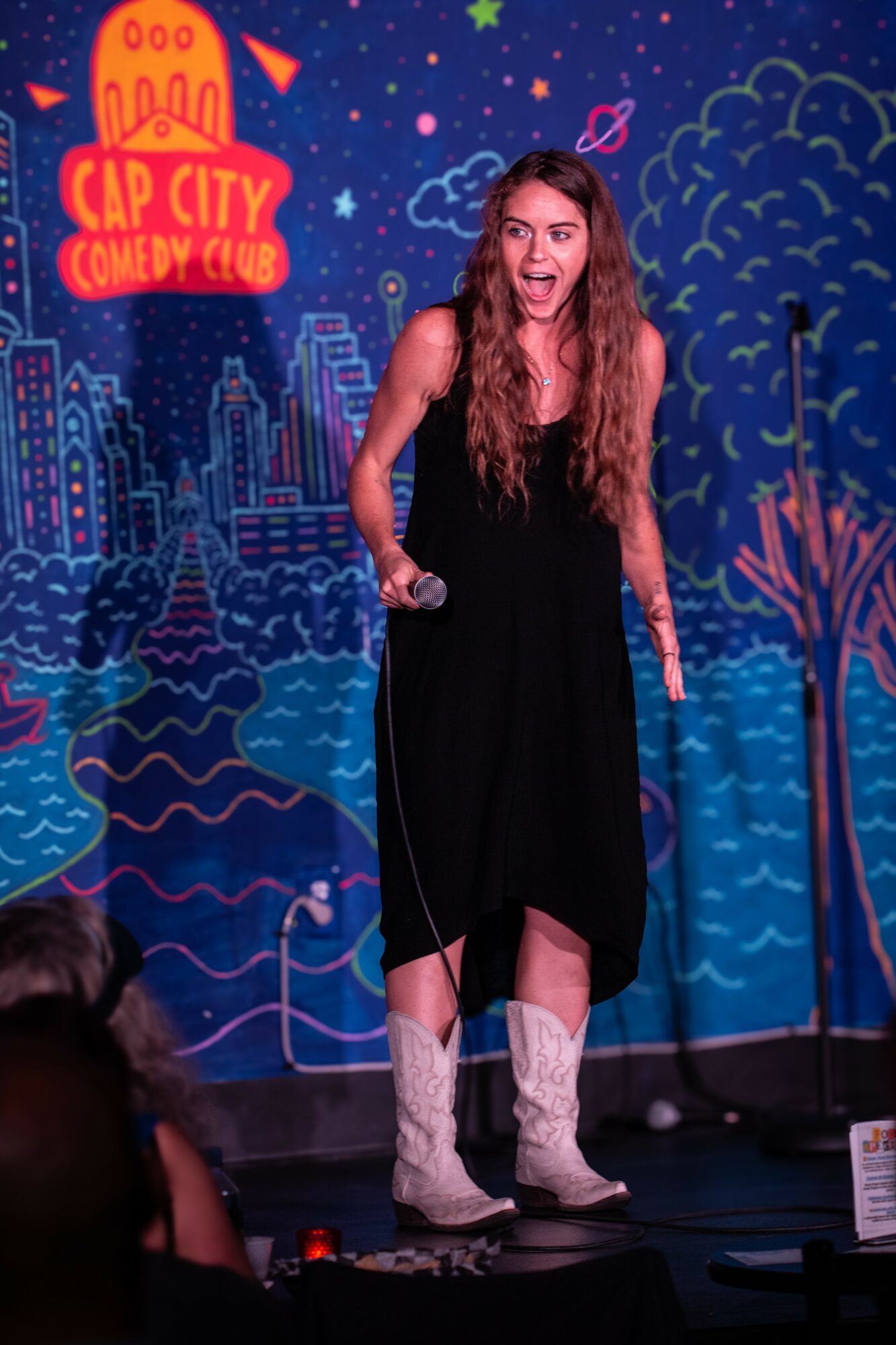 Woman performing stand-up comedy on stage with colorful cityscape backdrop, holding microphone, smiling, wearing a black dress and white boots.