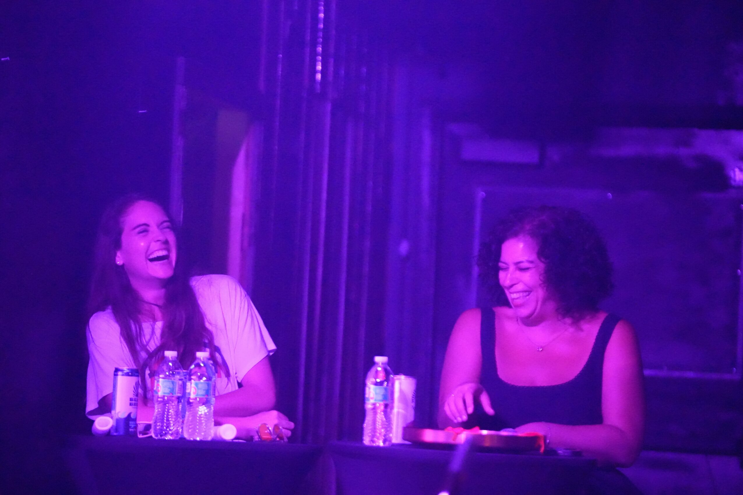 Two women sitting at a table, smiling and laughing, with water bottles in front of them, under purple lighting.