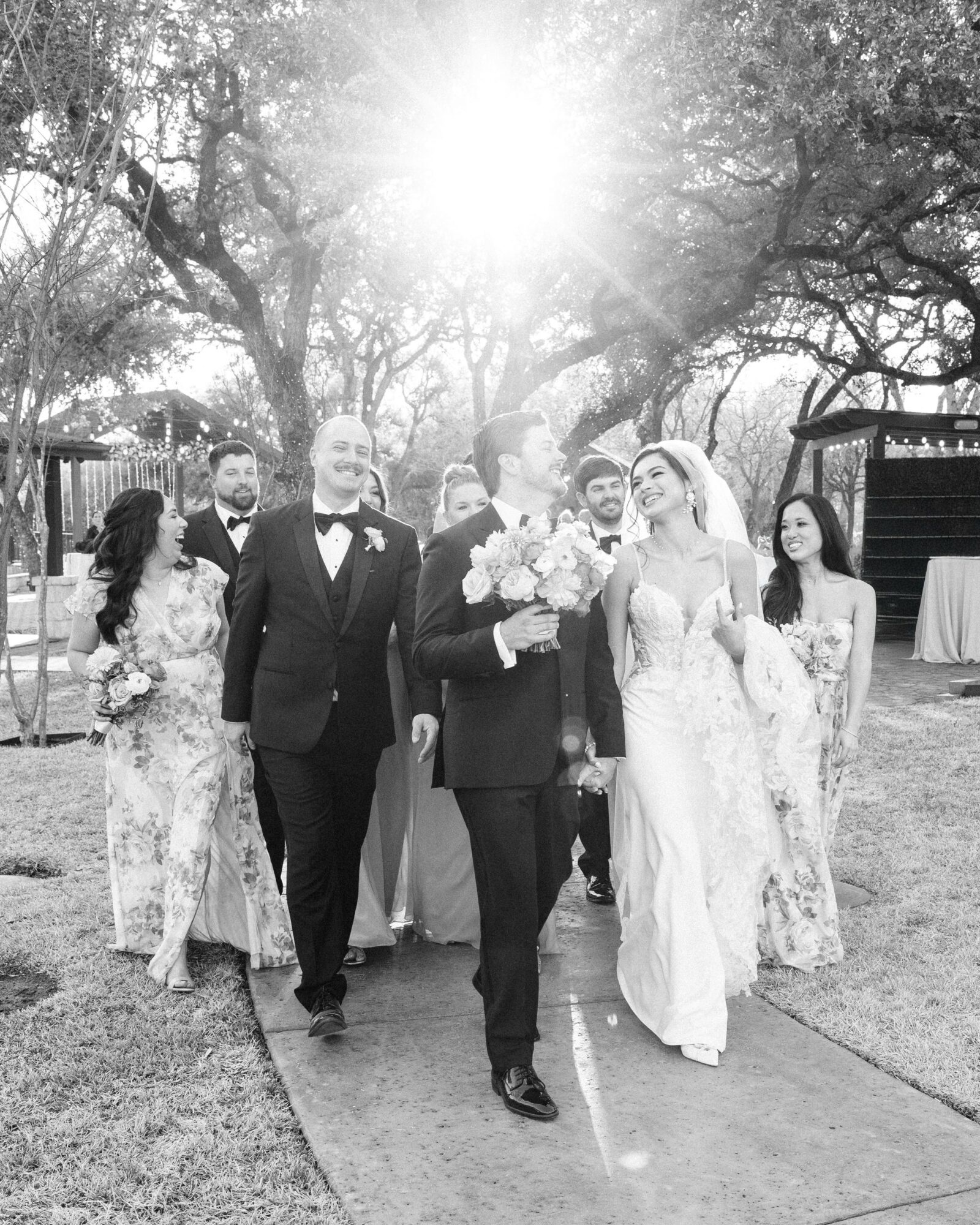 Group of people in wedding attire walking outdoors on a sunny day, trees in the background, some holding flowers.
