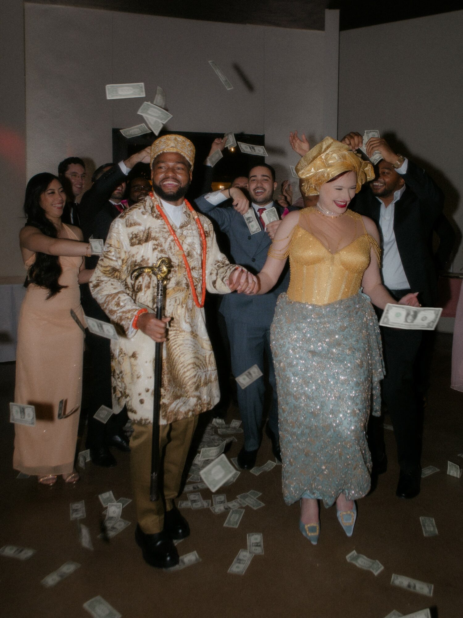 Group of people celebrating, some throwing money, with a man in traditional attire and a woman in a yellow dress in the foreground.