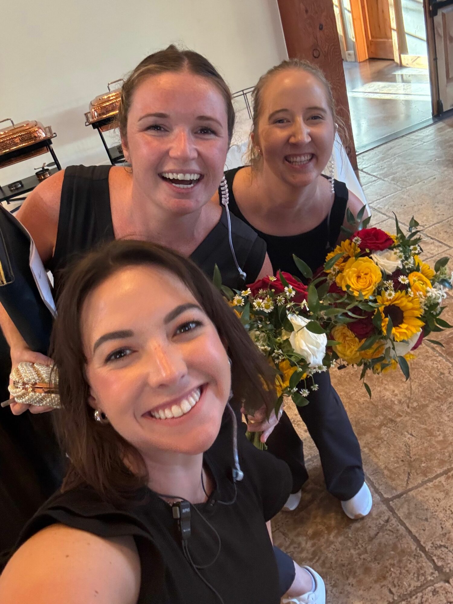Three women smiling, one holding a bouquet of yellow, white, and red flowers, in an indoor setting.