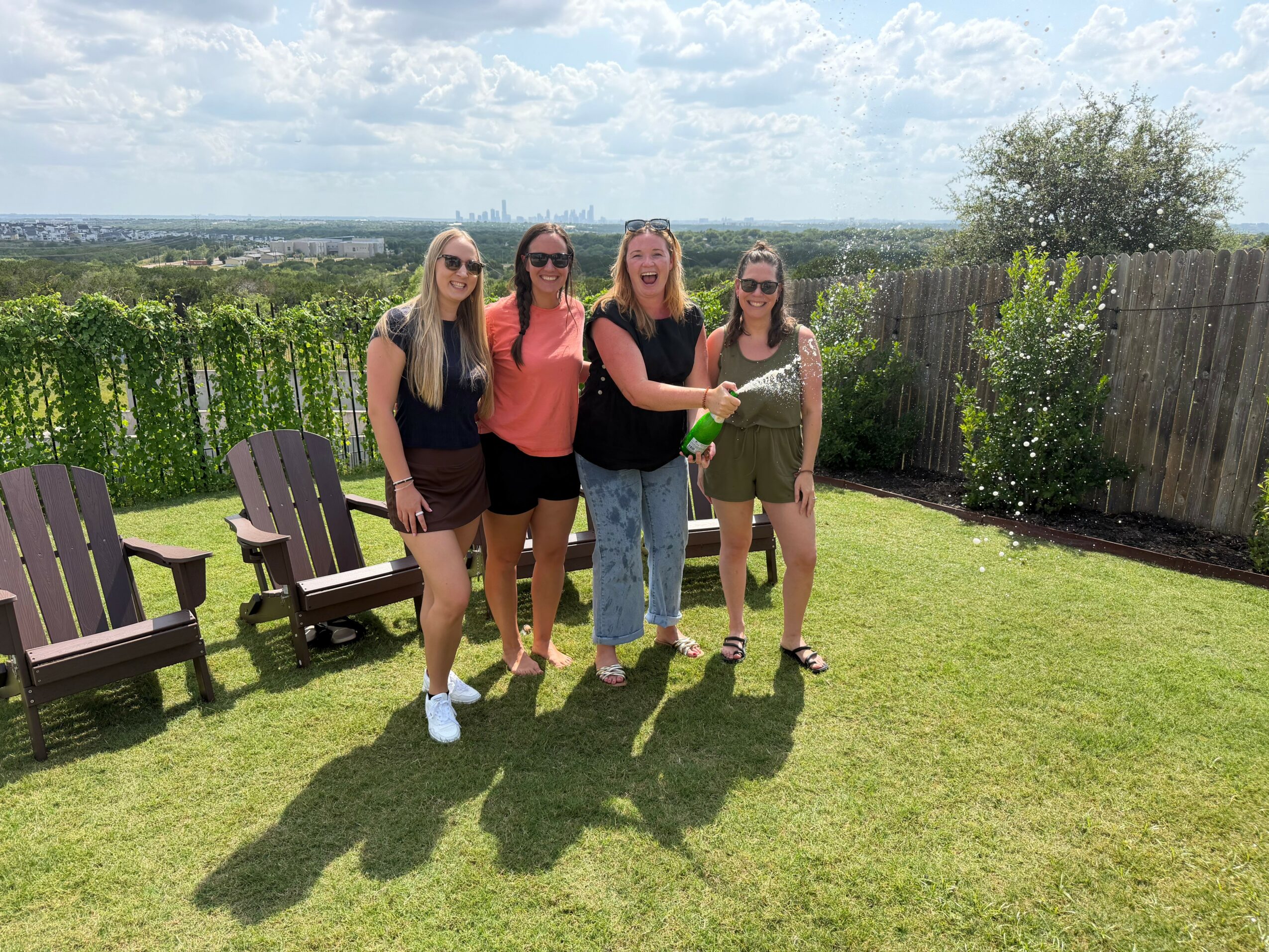 Four women standing on a grassy lawn with chairs, a fence, and trees in the background under a partly cloudy sky.