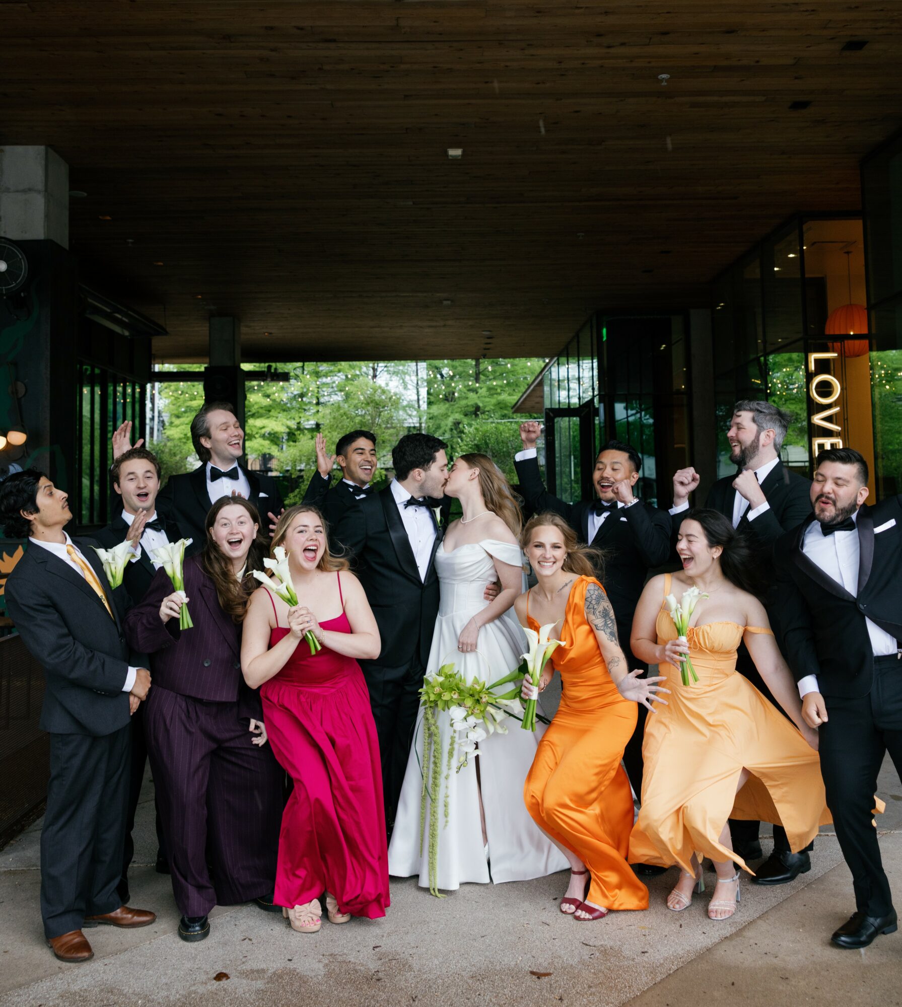 Group of people in formal attire celebrating outdoors, some dancing and smiling, with trees and a building in the background.