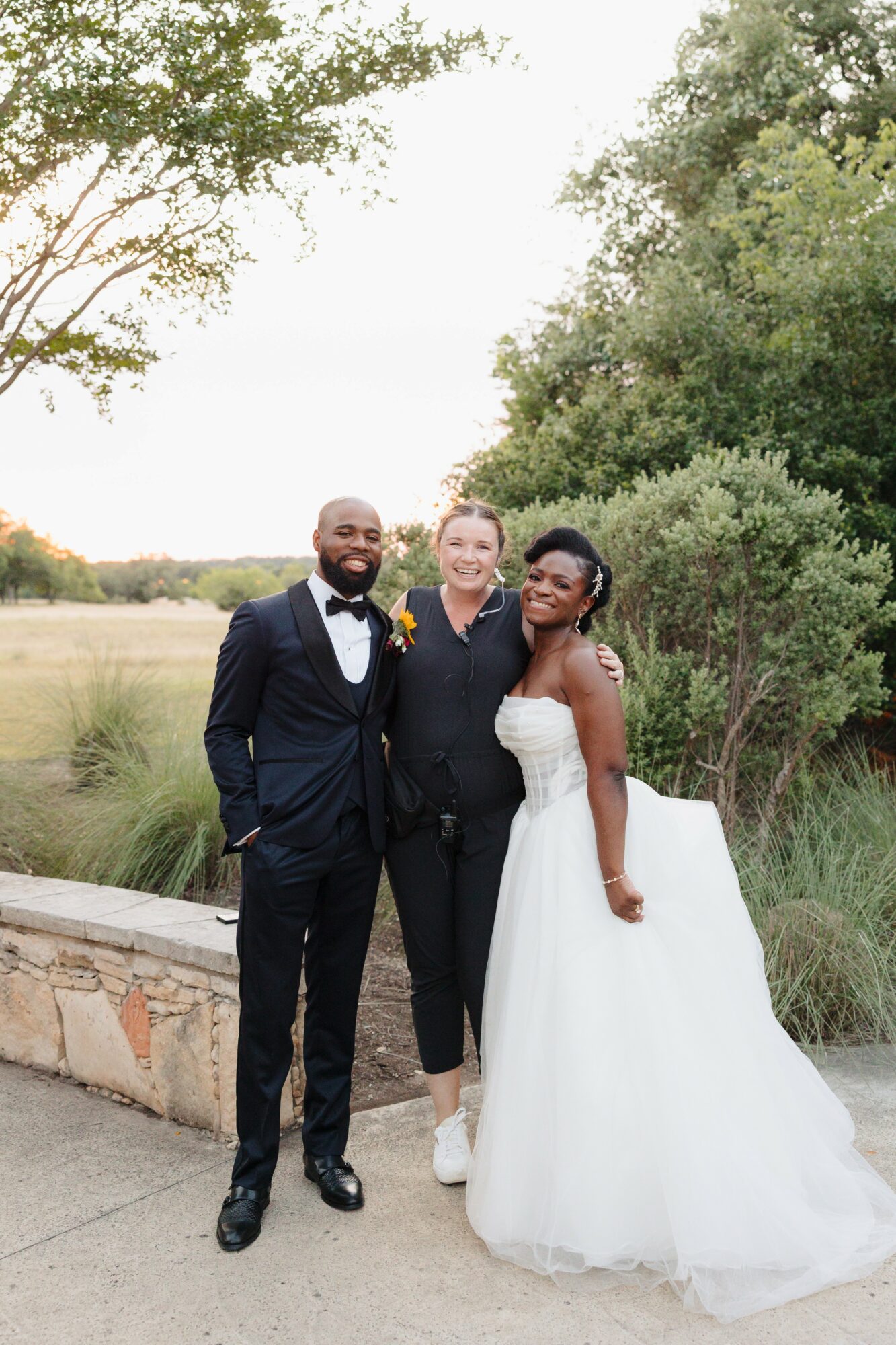 Three people standing outdoors, two women and one man, smiling, with trees and a grassy area in the background.