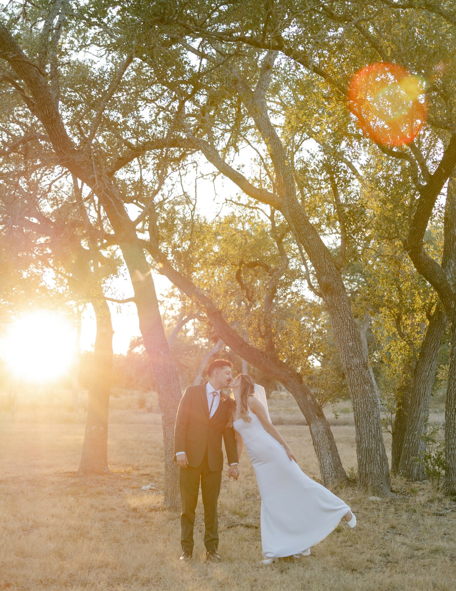 A couple in wedding attire standing outdoors among trees with sunlight shining through branches.