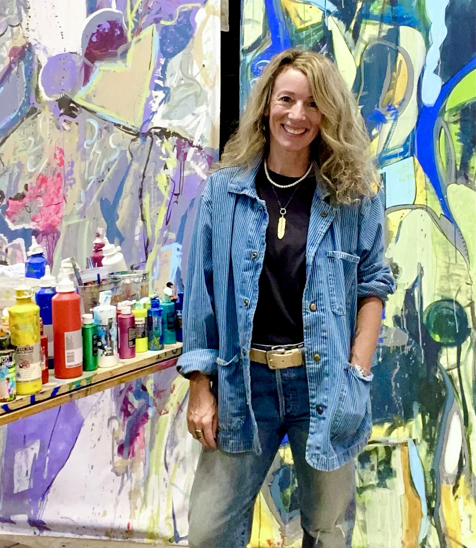 Woman with blonde hair smiling, wearing a denim jacket and black shirt, standing in front of colorful abstract art, with art supplies on a table.