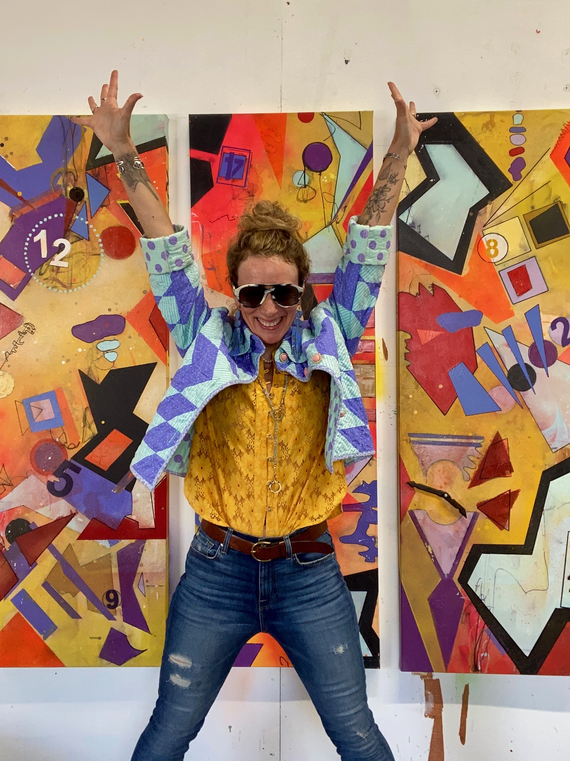 Person with sunglasses and curly hair, smiling with arms raised, standing in front of colorful abstract artwork.