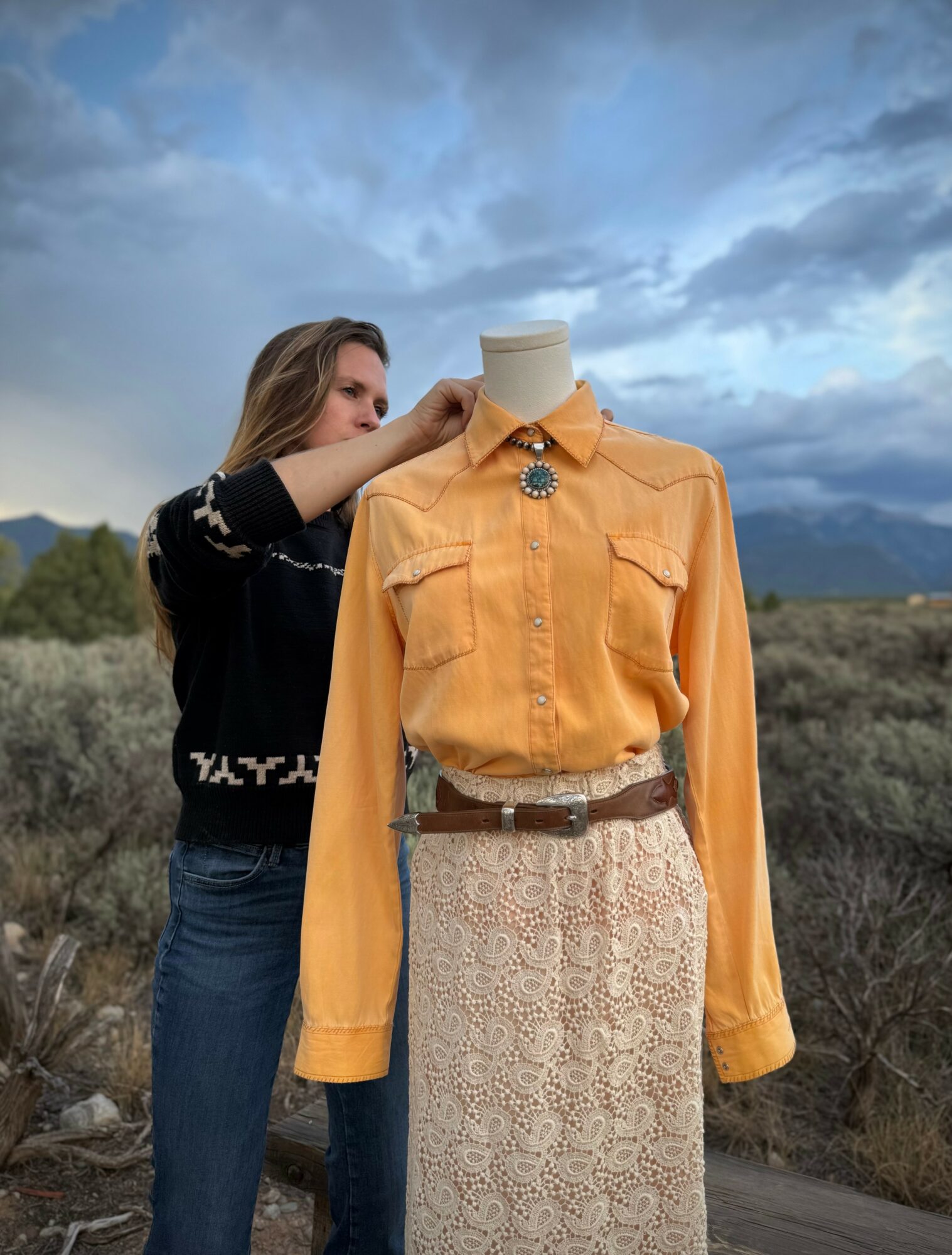 Person adjusting a mannequin dressed in an orange shirt and lace skirt outdoors, cloudy sky background.