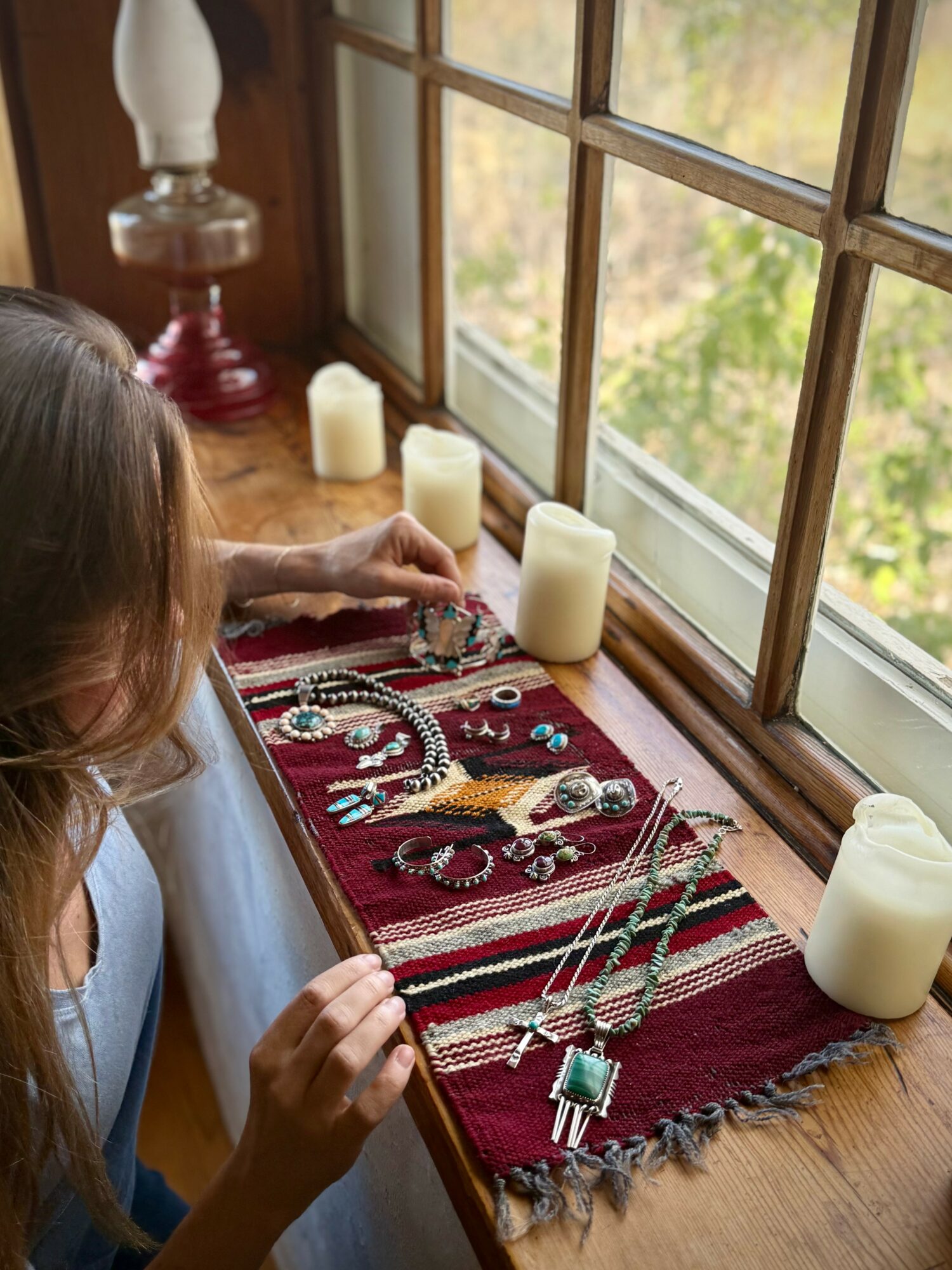 Person arranging jewelry on a red cloth by a window with candles, outdoors visible through the window.