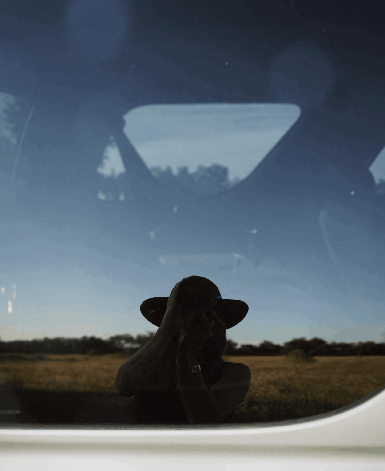Person taking a photo in a vehicle window, with a landscape and sky visible outside, reflection in the glass.