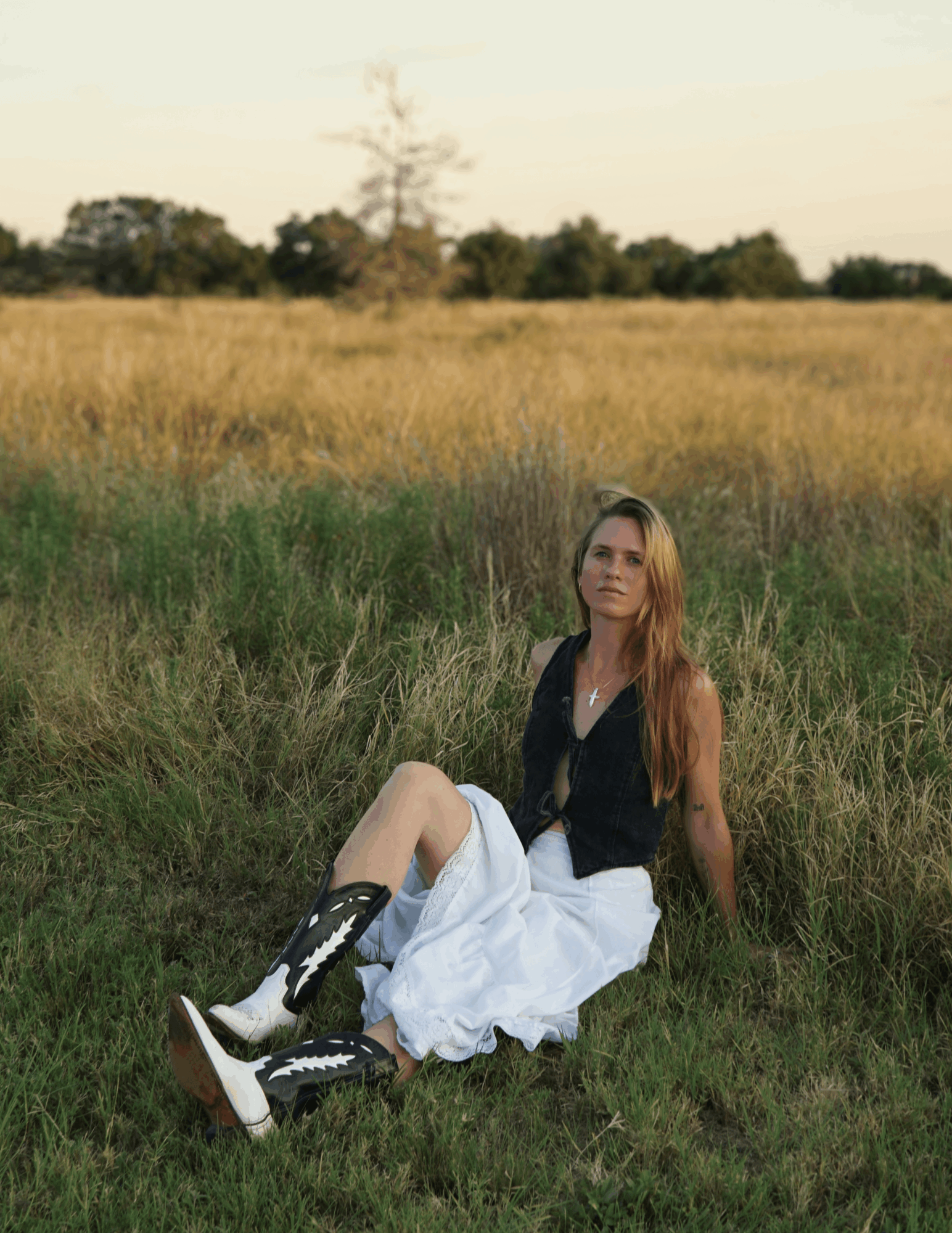 Woman with long hair sitting on grass in a field, wearing a black top, white skirt, and boots, looking at the camera.