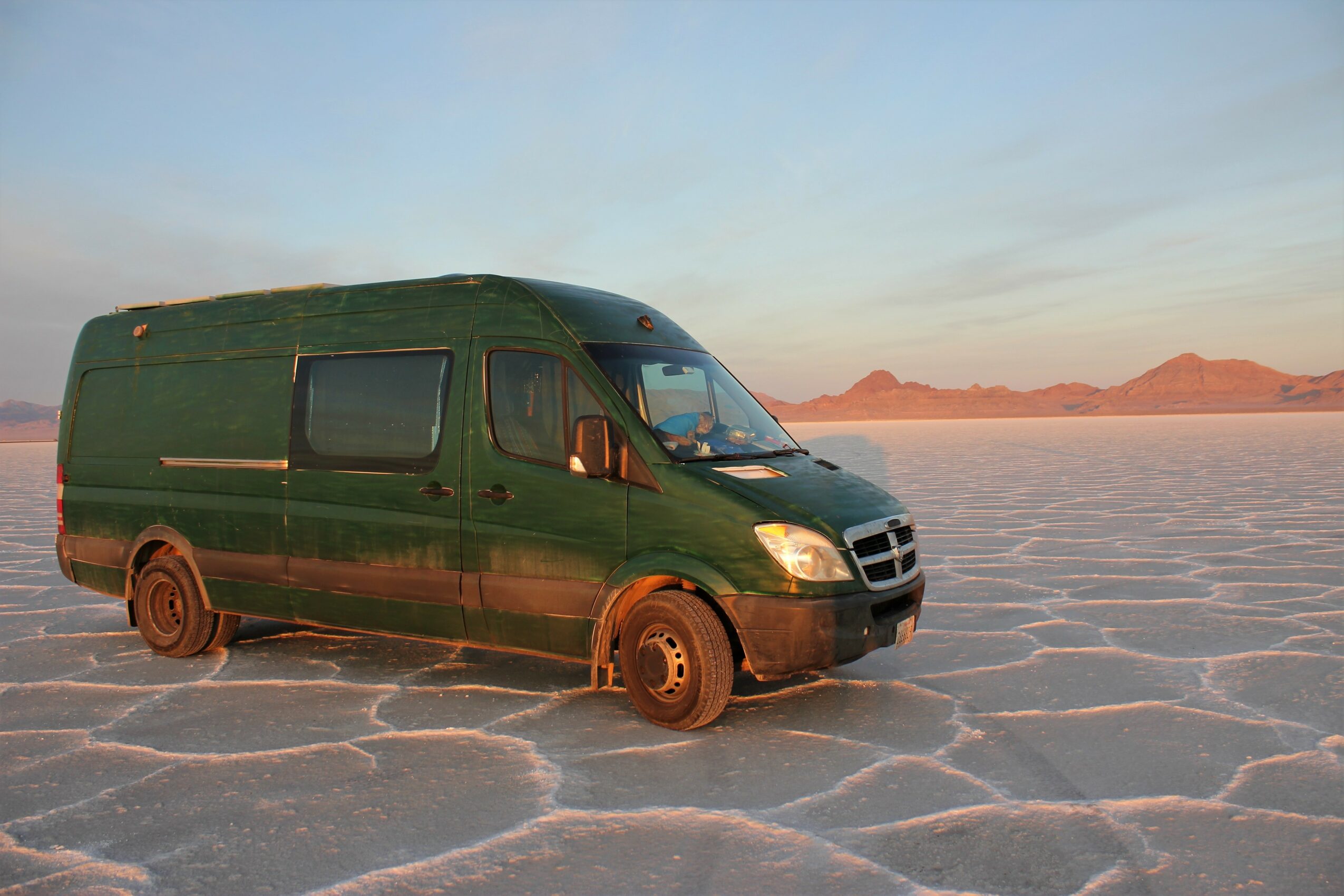 Green van partially buried in salt flats with mountains in the background, under a clear sky.