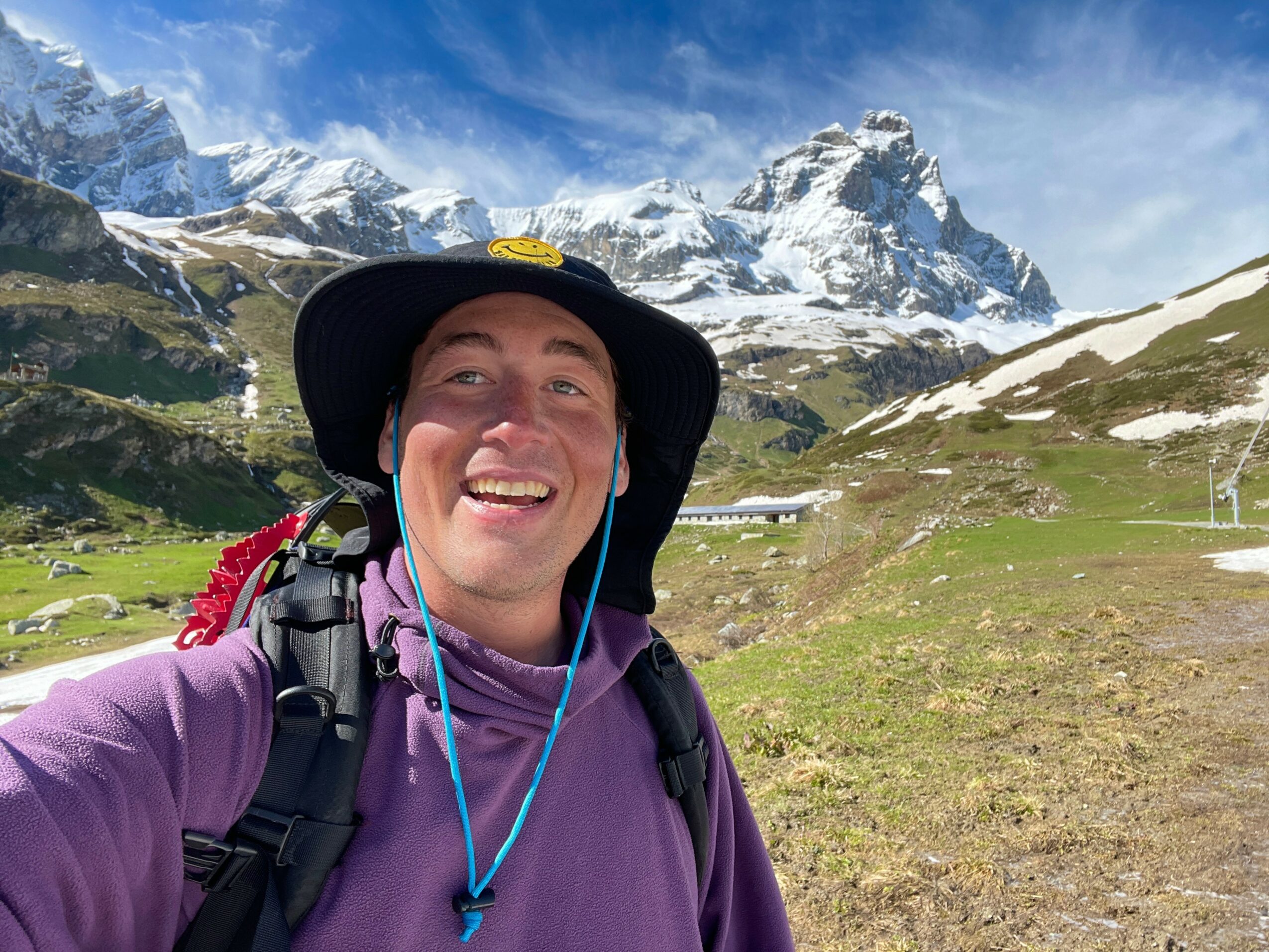 Person smiling outdoors with snow-capped mountains and green landscape in background.