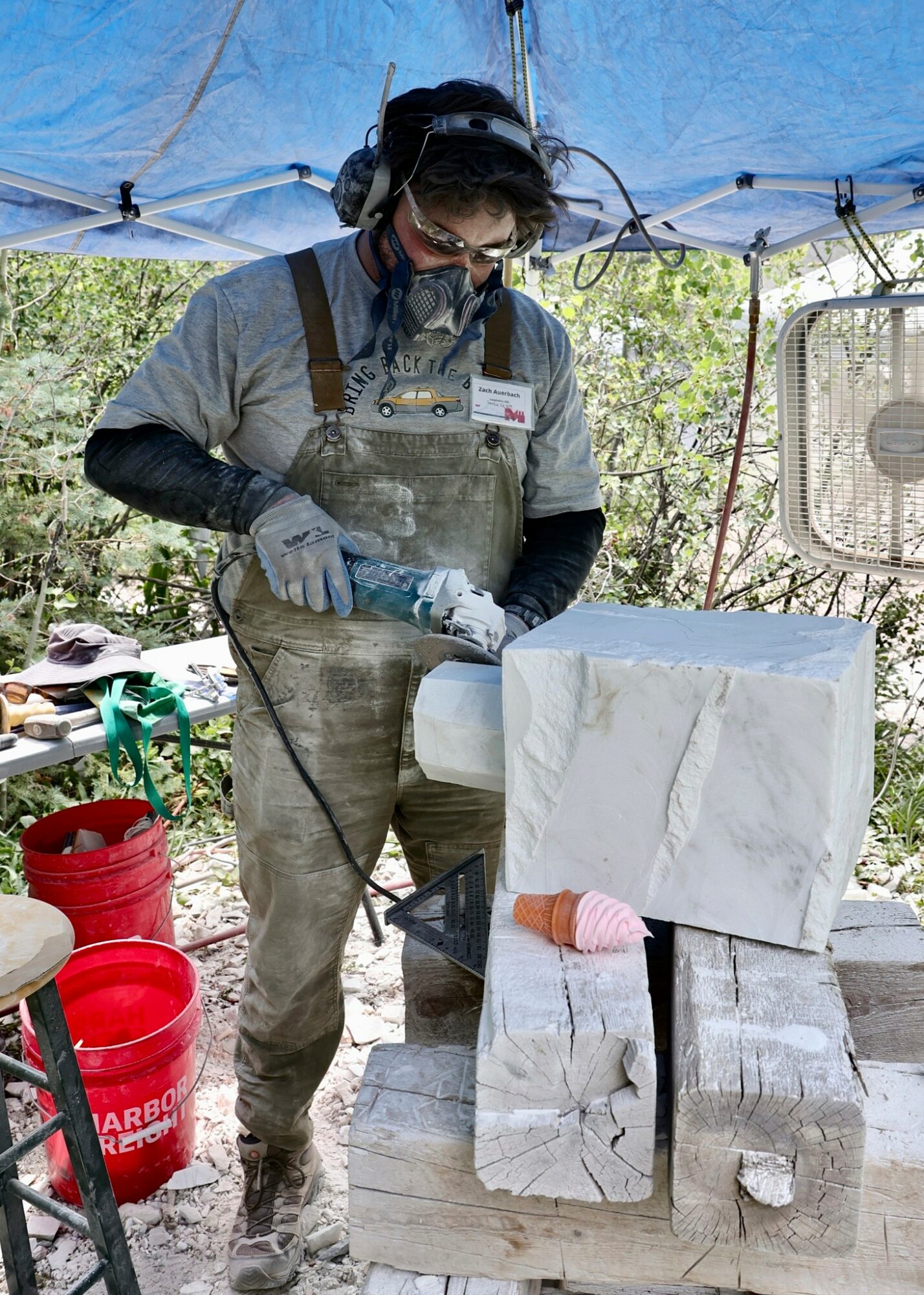 Person wearing protective gear carves a block of stone outdoors under a blue canopy, surrounded by trees and equipment.
