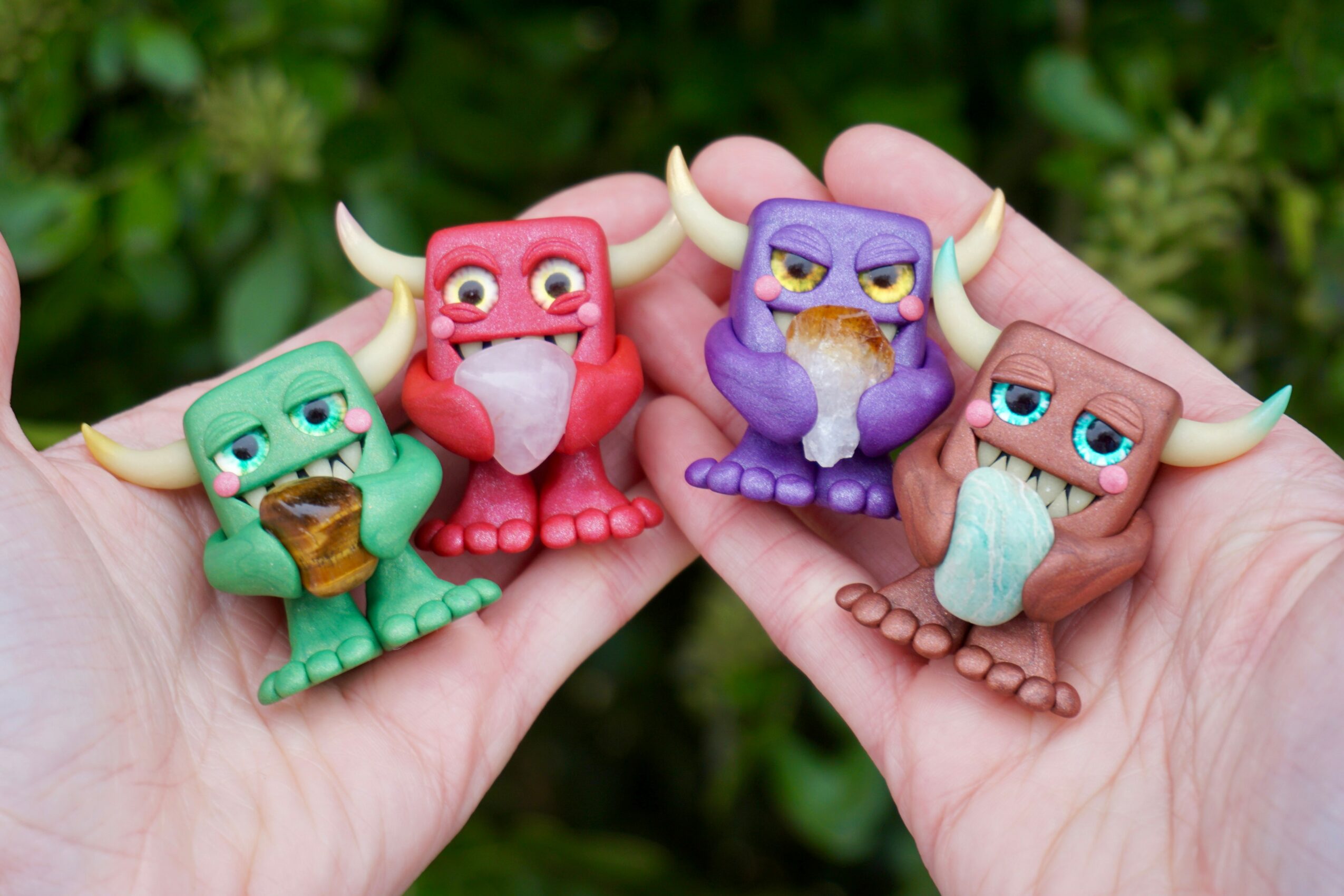 Four small colorful dragon figurines held in hands outdoors, each with different colors and expressions, against green foliage background.