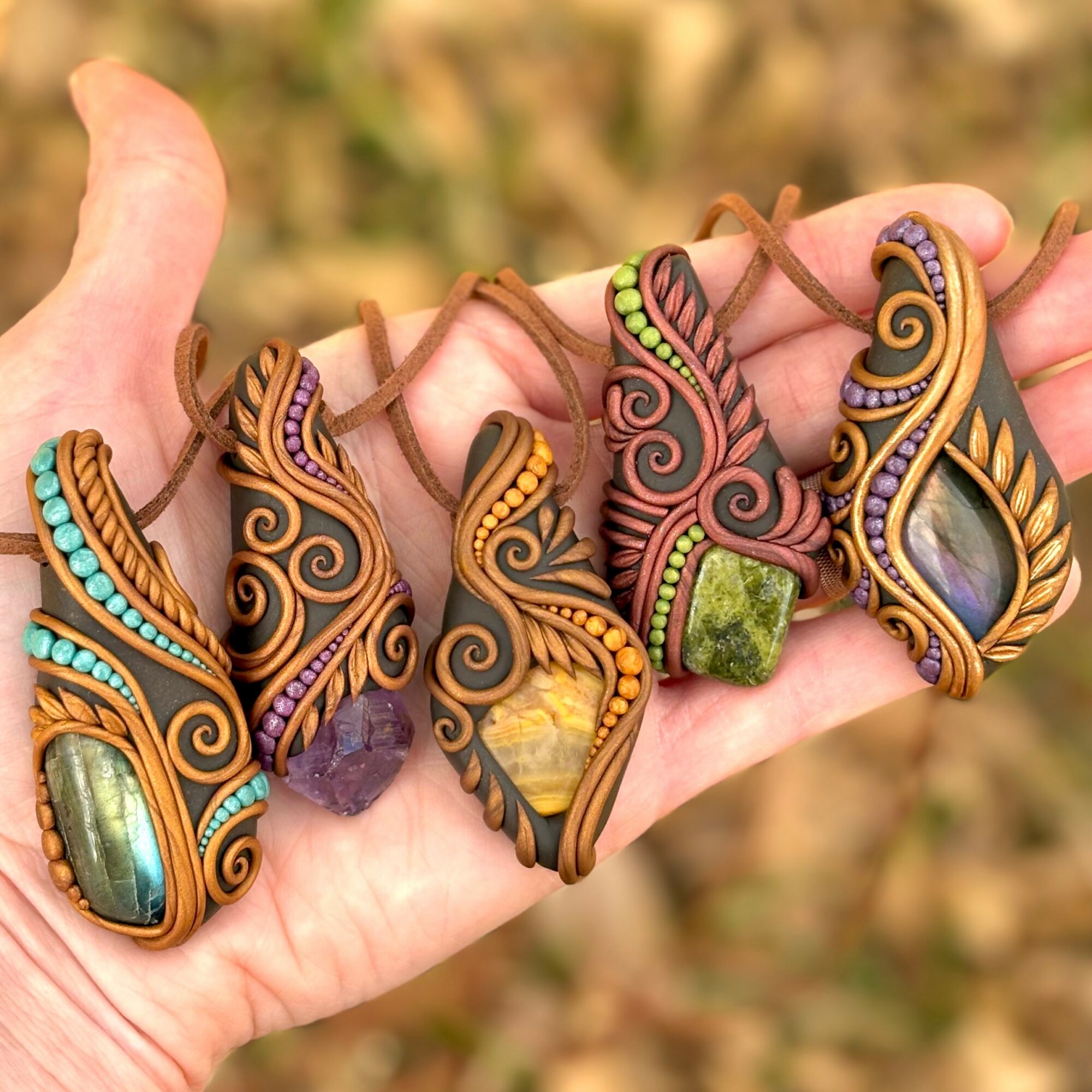 Hand holding five colorful, intricately designed pendants with stones and swirling patterns, outdoors background.