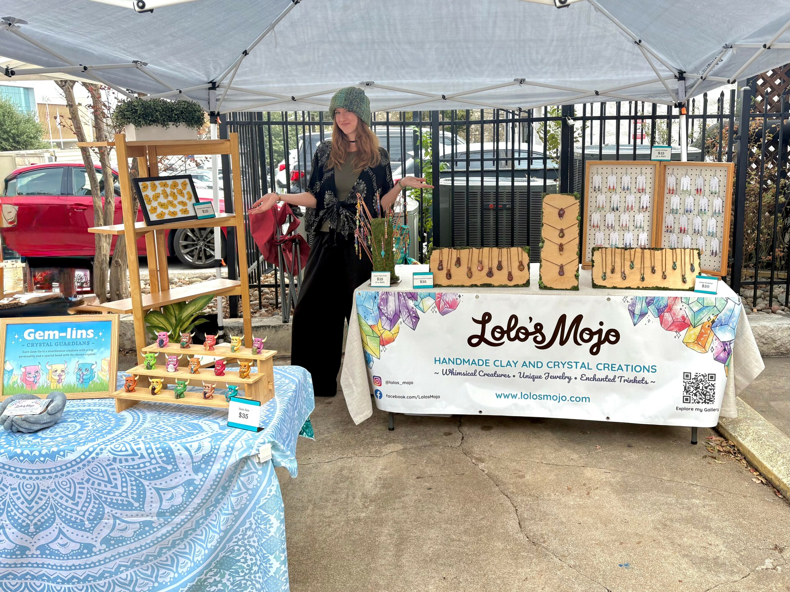 Woman standing behind a table with handmade clay and crystal creations, outdoors under a canopy, with a fence and parked cars in background.