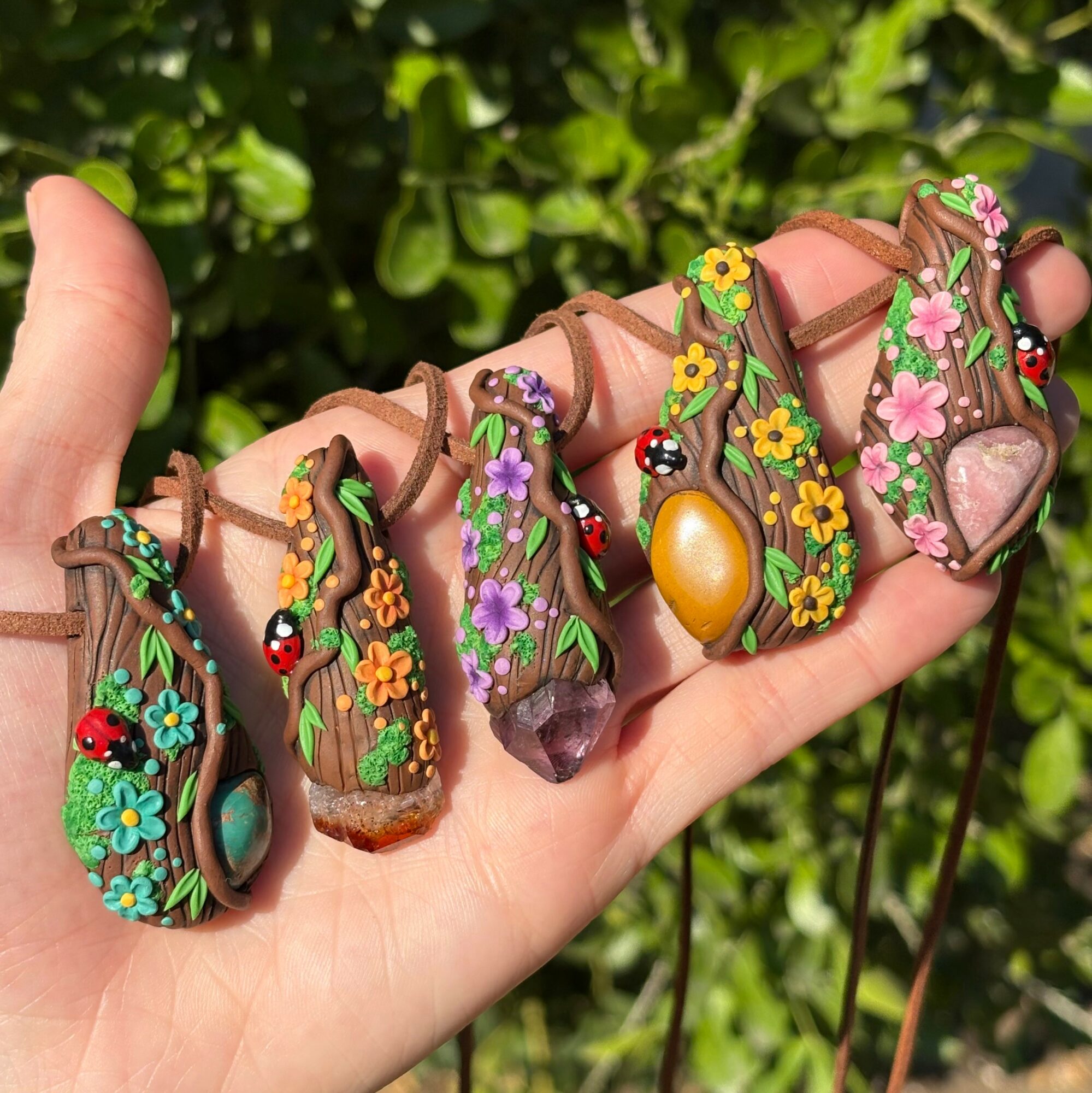 Hand holding five colorful, decorated pendants resembling tree trunks with flowers, ladybugs, and gemstones, outdoors with green foliage background.