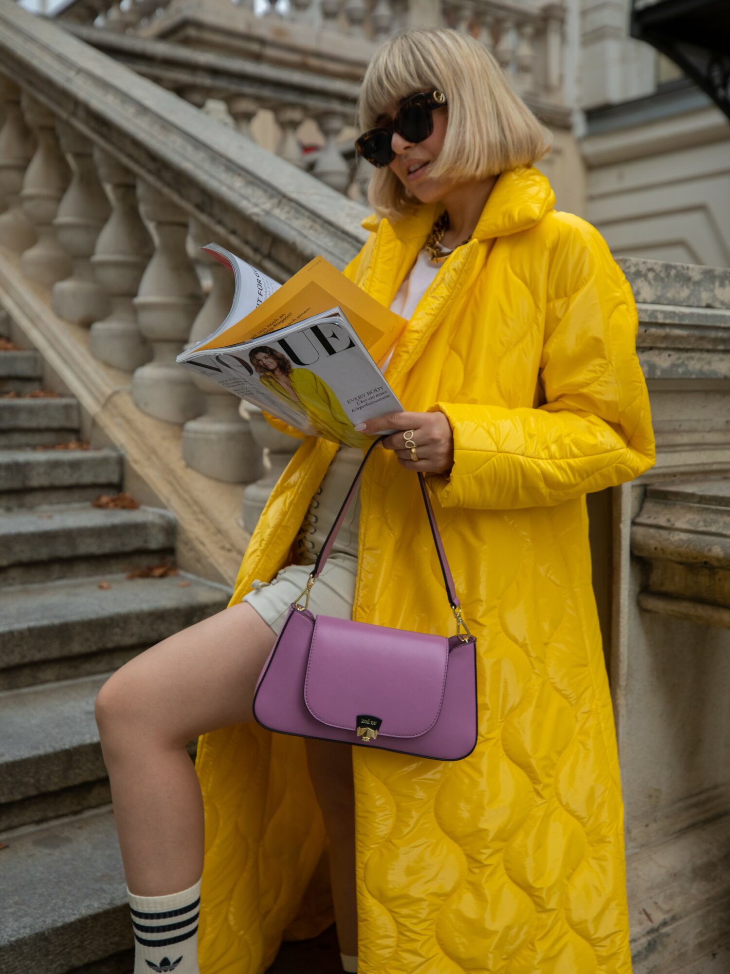 Woman in yellow coat reading a magazine, carrying a purple purse, sitting on stairs outside building.
