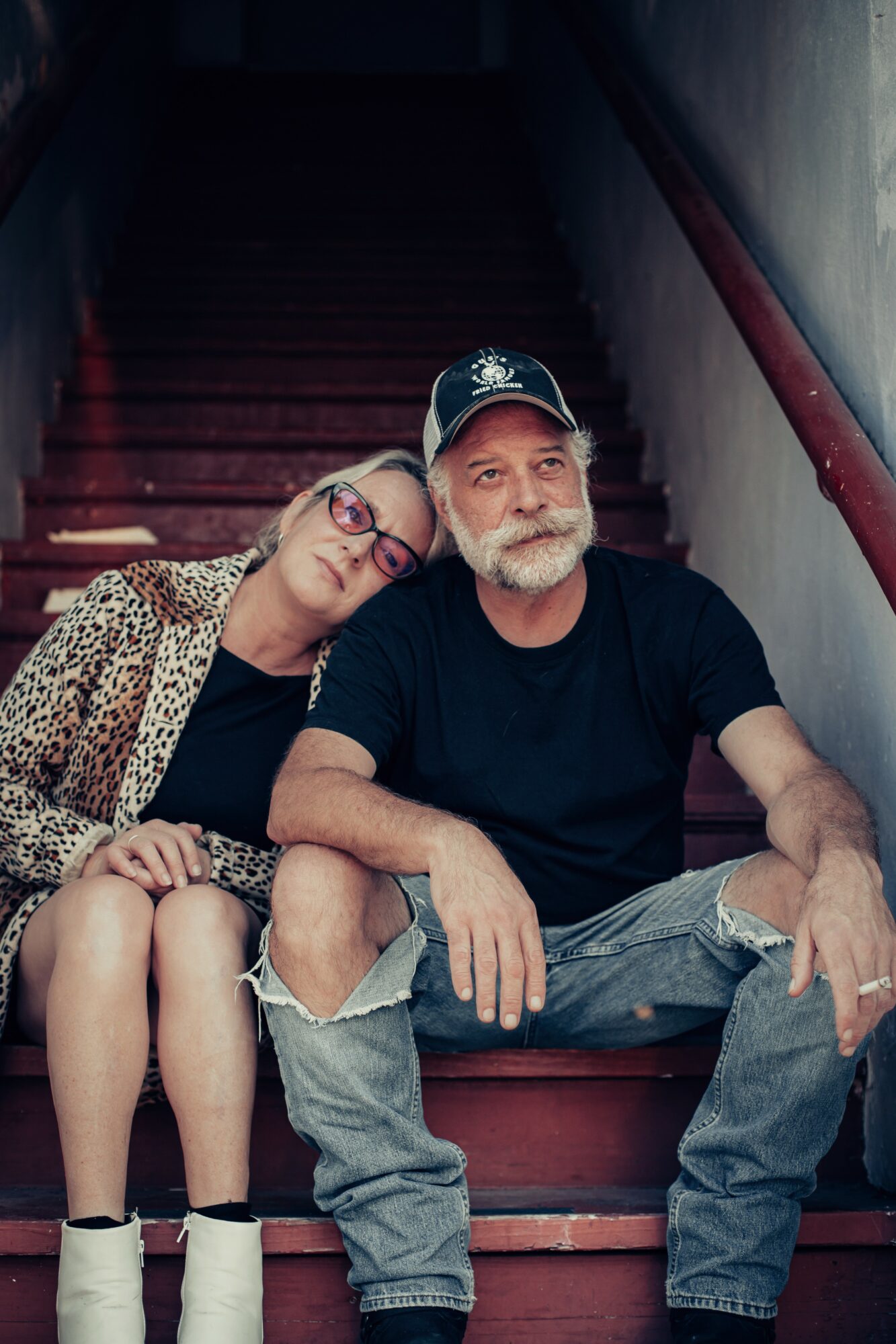 A woman with glasses and a man with a beard sit on stairs, leaning on each other, in a dimly lit setting.
