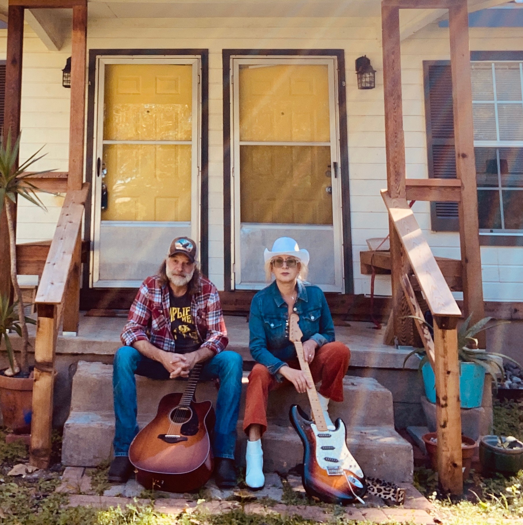 Two people sit on front steps with guitars, in front of yellow doors, wooden porch, and house exterior, smiling.