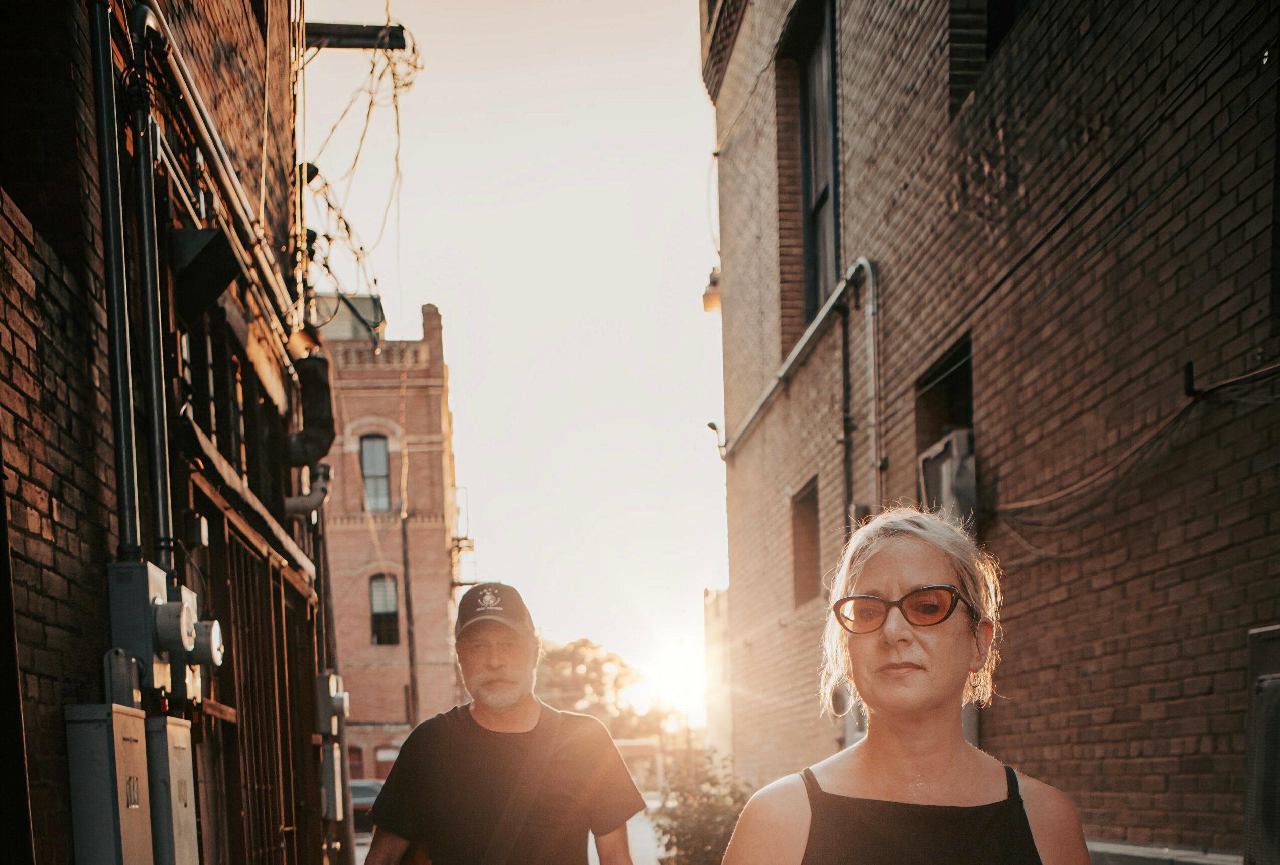 Two women walking in an alleyway with brick buildings, sunlight shining behind them, one wearing sunglasses.