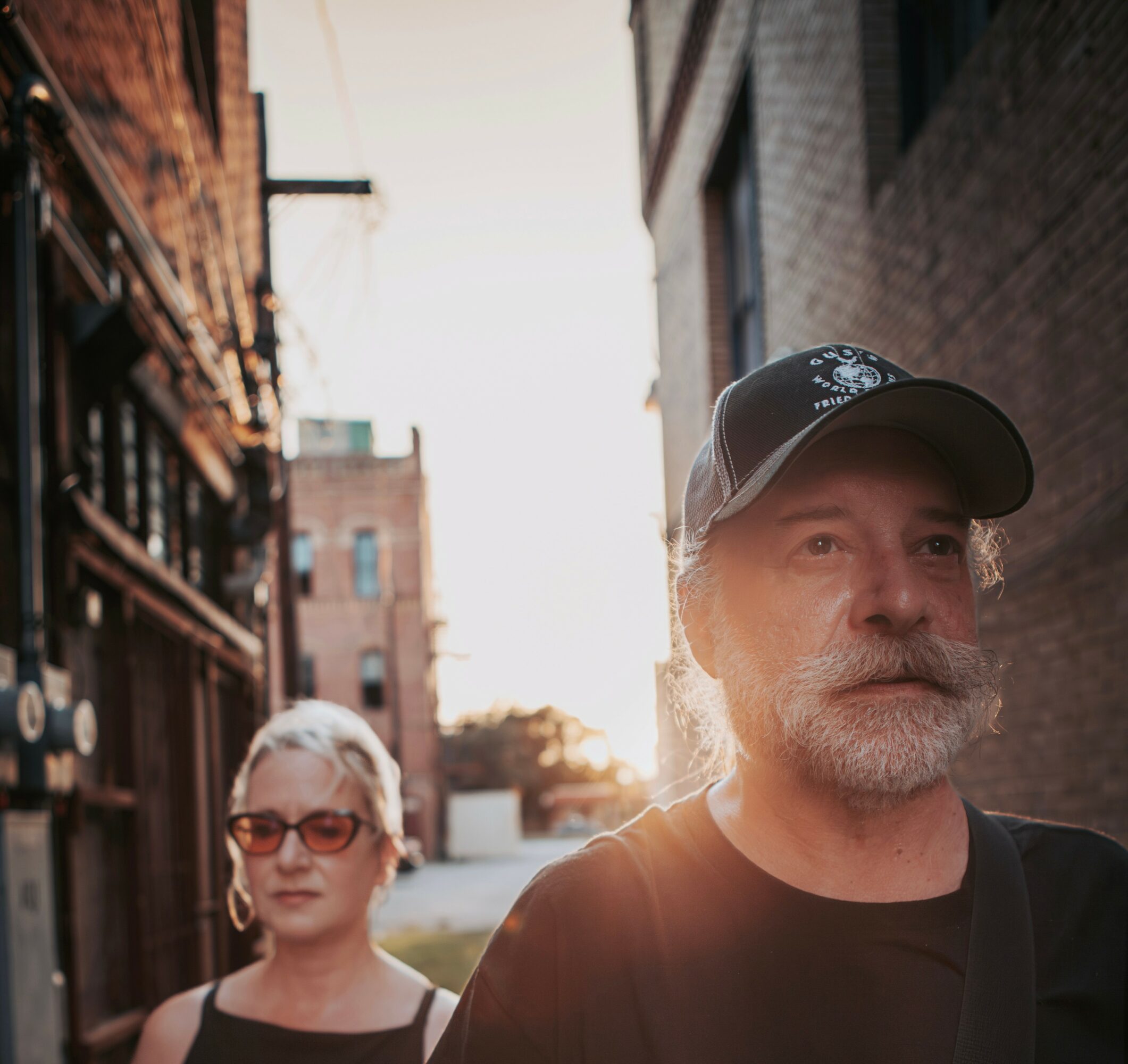 A man with a beard and a woman with glasses walking outdoors in an urban setting during sunset.