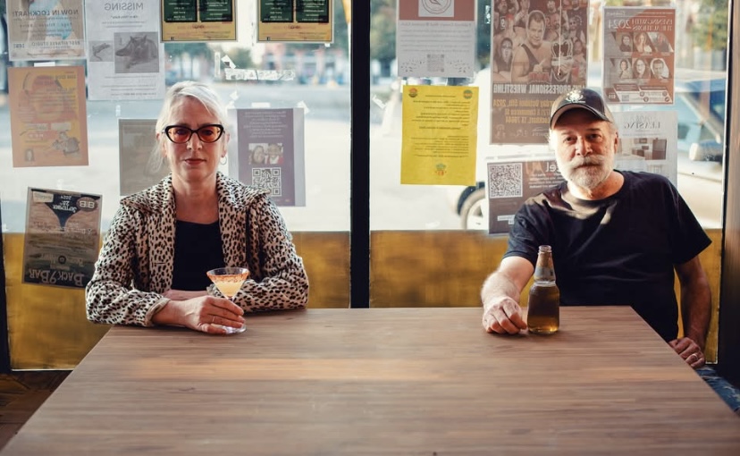 Two people sitting at a wooden table in a cafe, with posters on the window behind them.