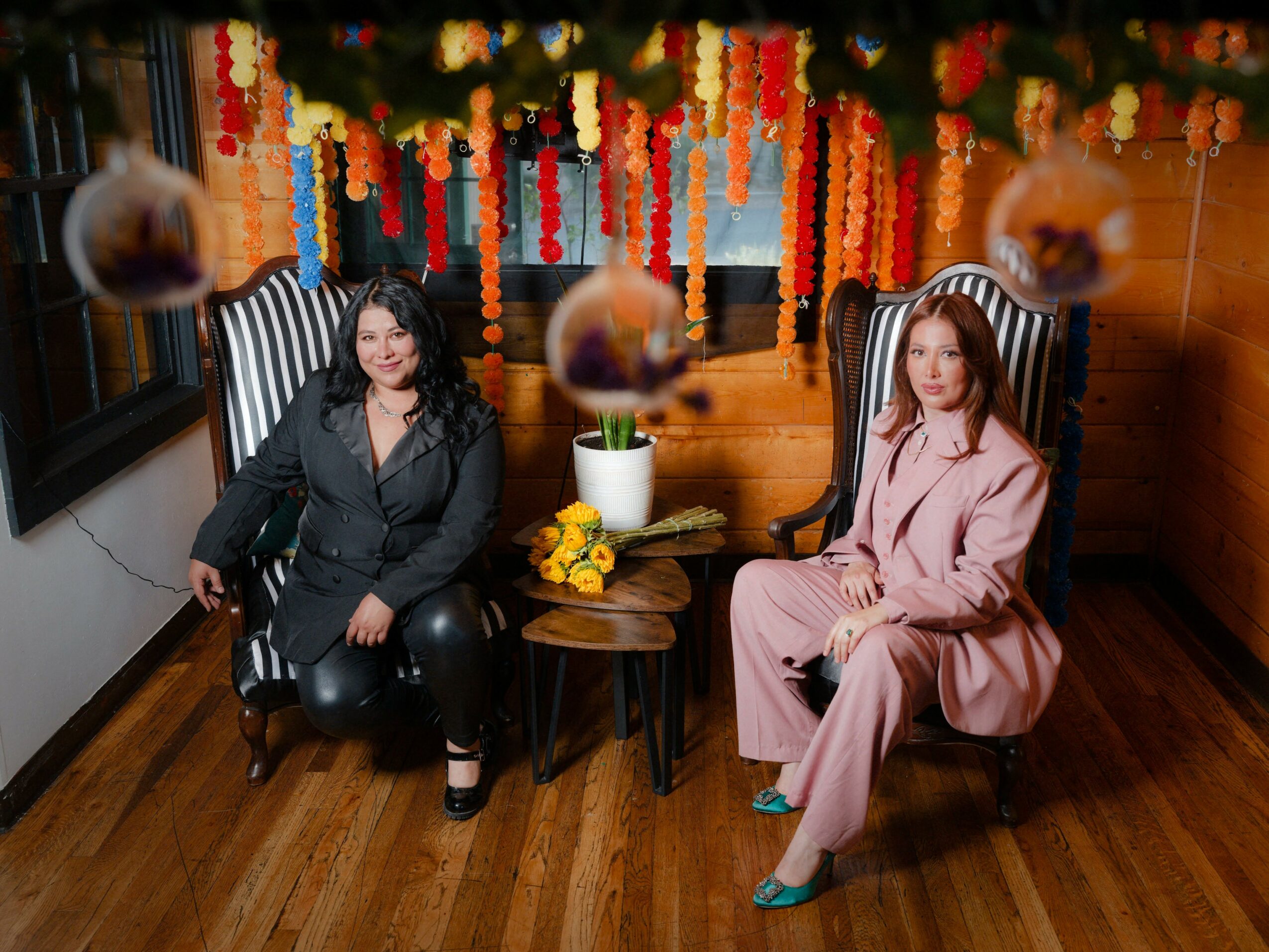 Two women sit in high-backed chairs with a wooden wall and colorful hanging decorations behind them. A small table with a potted plant and flowers is between them.