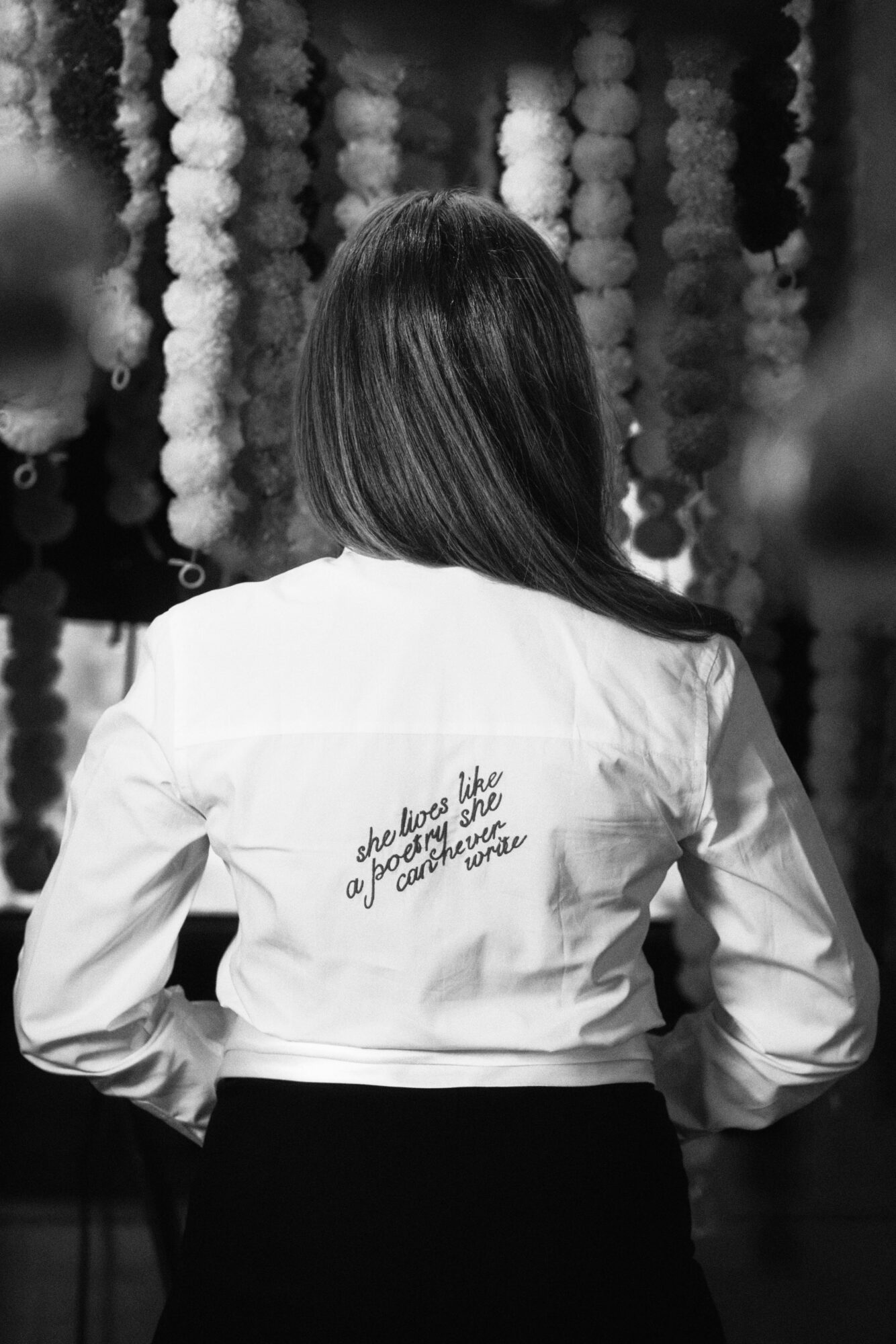 Back of a woman with long hair wearing a white jacket with text on the back, in front of hanging decorations.