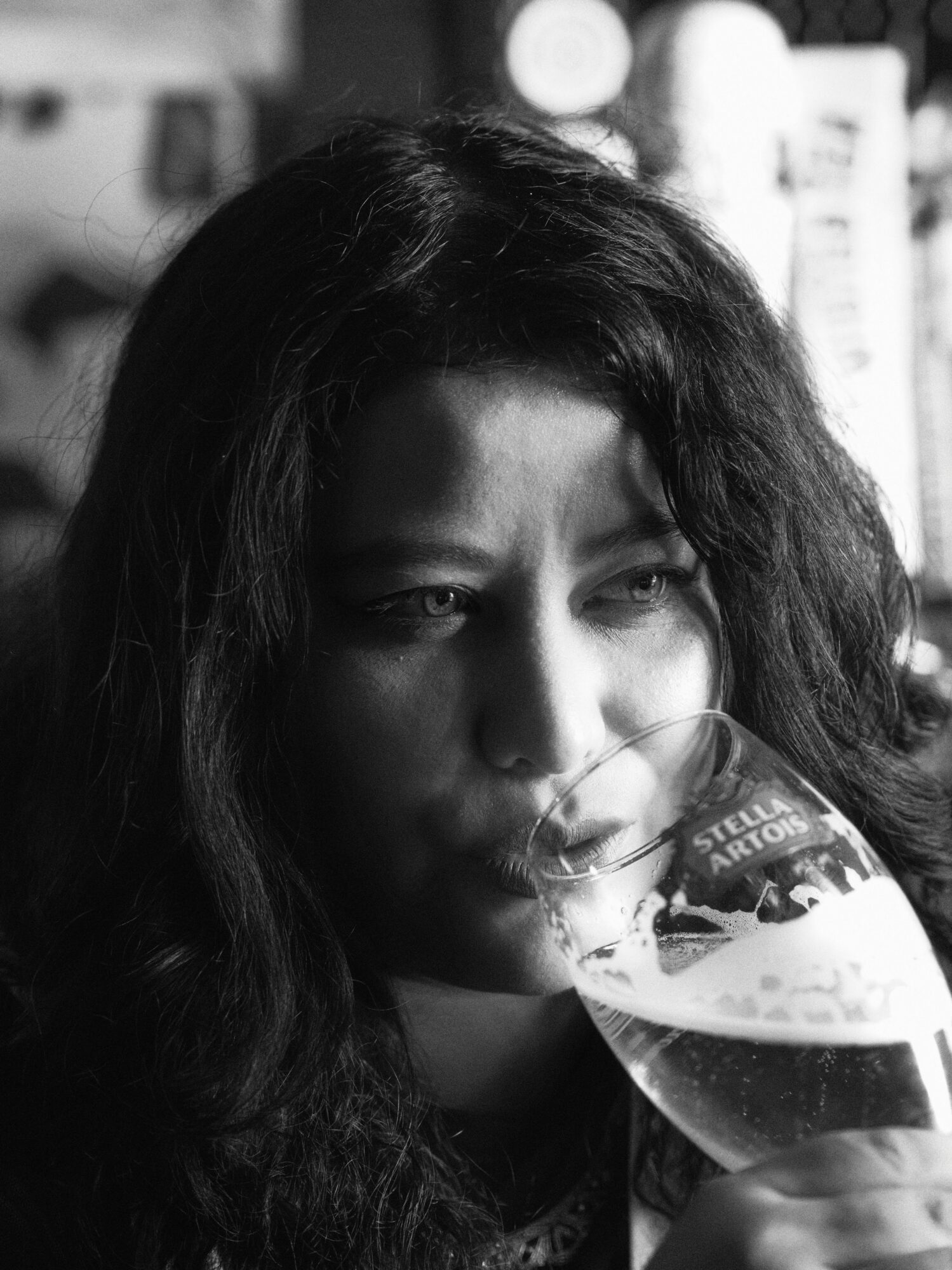 Woman with long dark hair drinking from a glass, in black and white, with blurred background.