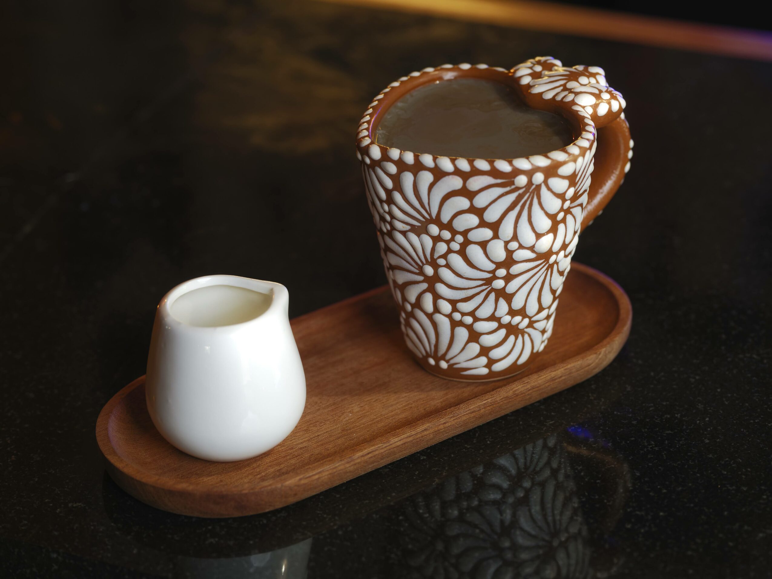 A decorated mug filled with a hot beverage and a small white pitcher on a wooden tray, on a dark surface.