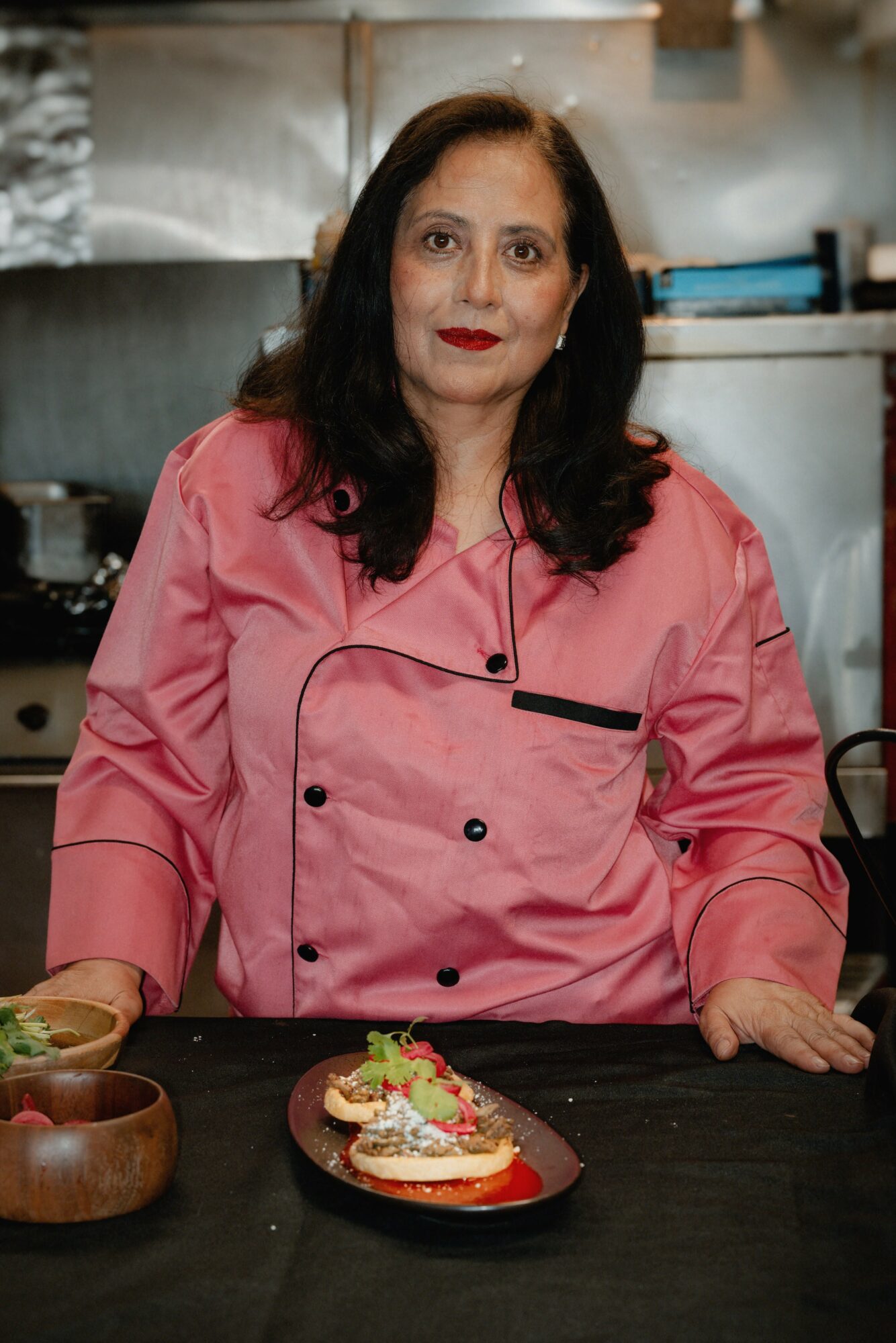 Woman in pink chef's coat standing behind a table with a decorated dish, kitchen background, smiling, dark hair, red lipstick