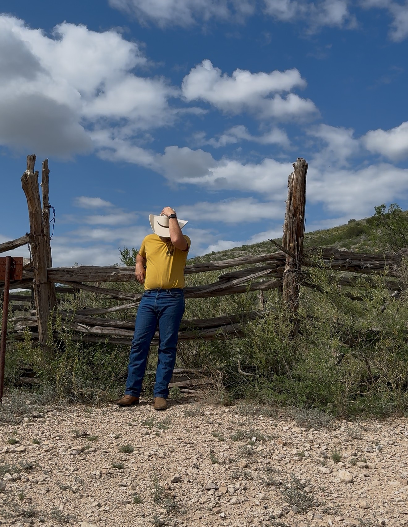 Person wearing a yellow shirt and cowboy hat standing in front of a wooden fence outdoors under a partly cloudy sky.