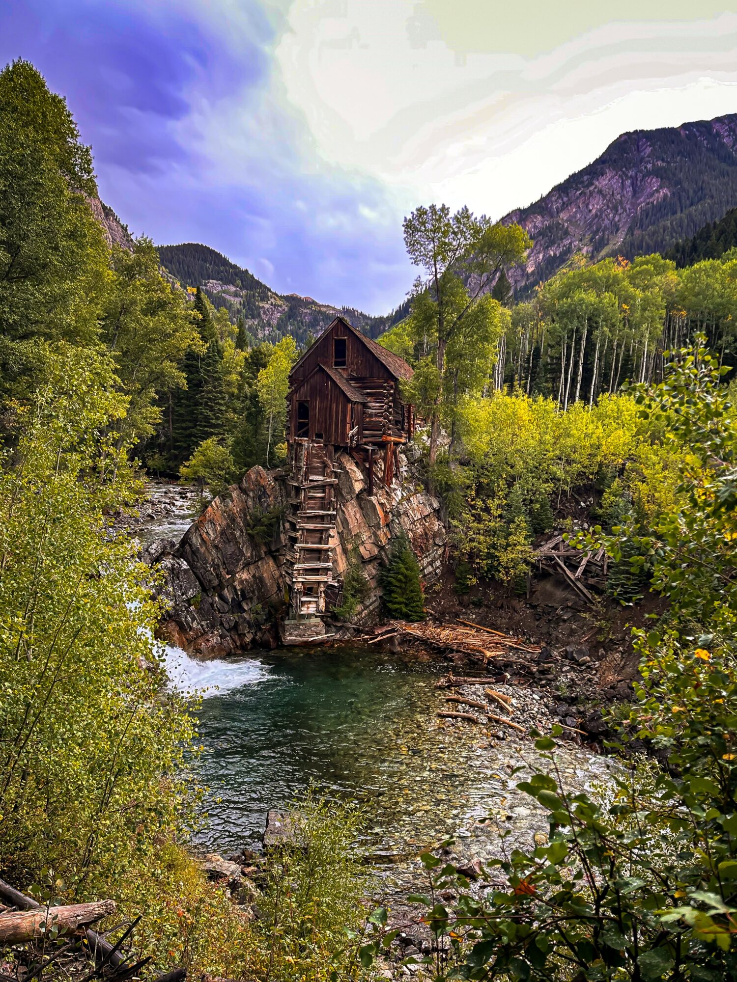 Wooden house on a rocky cliff above a river, surrounded by green trees and mountains, under a partly cloudy sky.