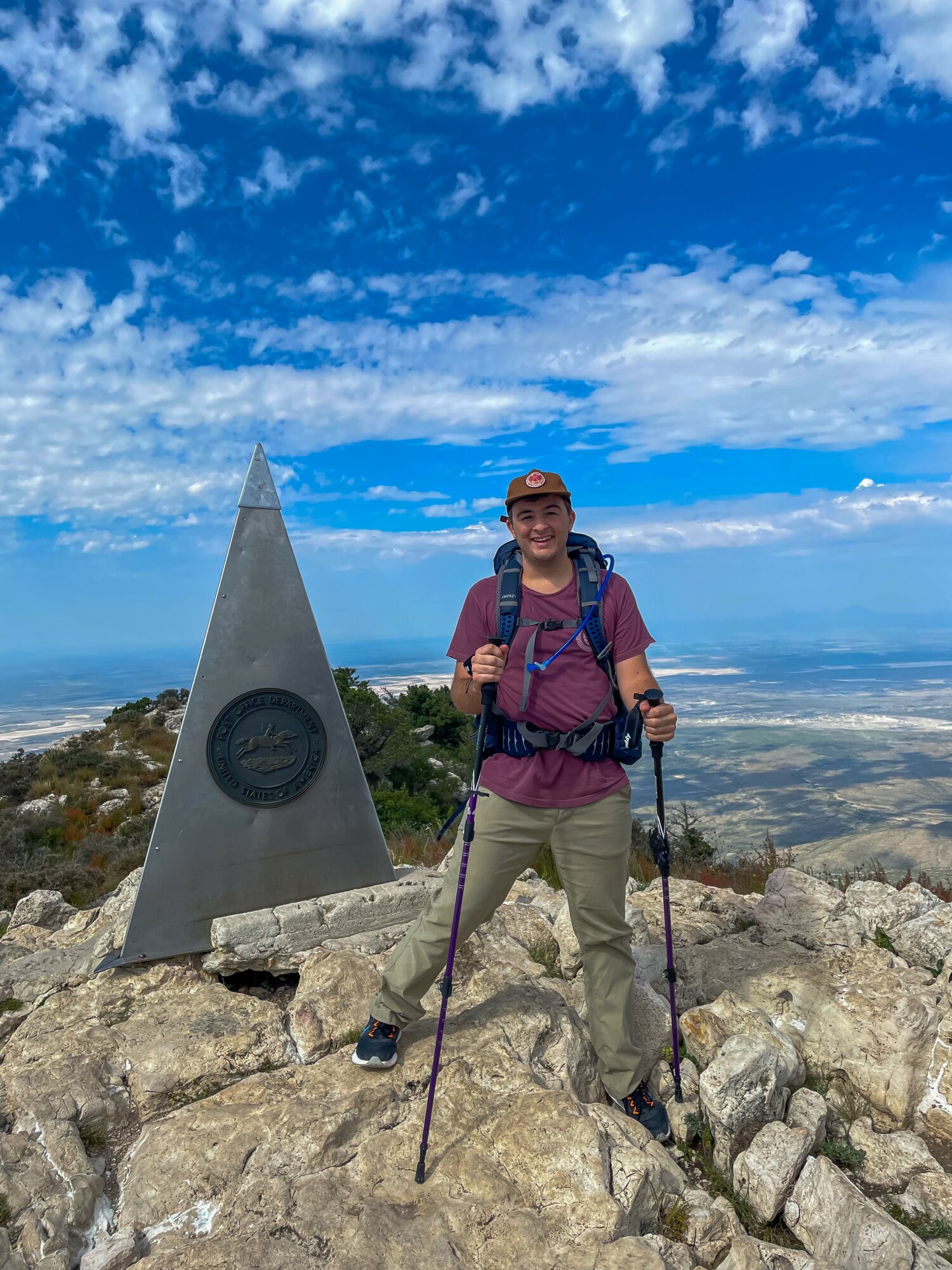 Person standing on rocky mountain summit with a triangular marker, blue sky with clouds in background.
