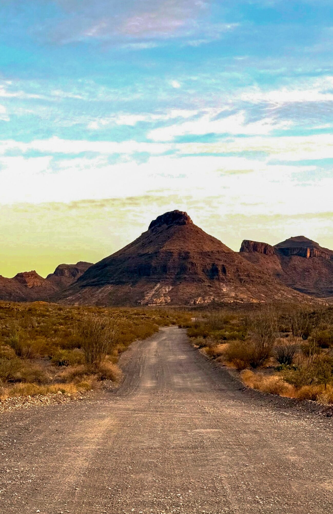 Dirt road leading to mountain with layered rock formations under a partly cloudy sky.