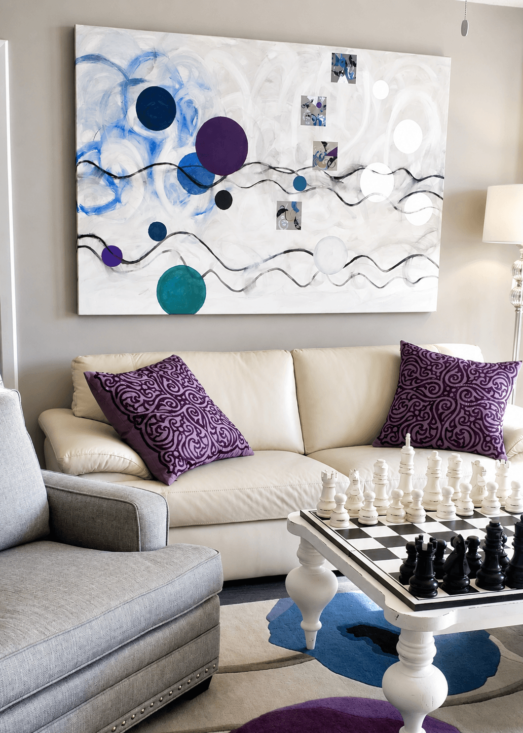 Living room with a white sofa, purple pillows, a chessboard, and an abstract painting with circles and wavy lines on the wall.