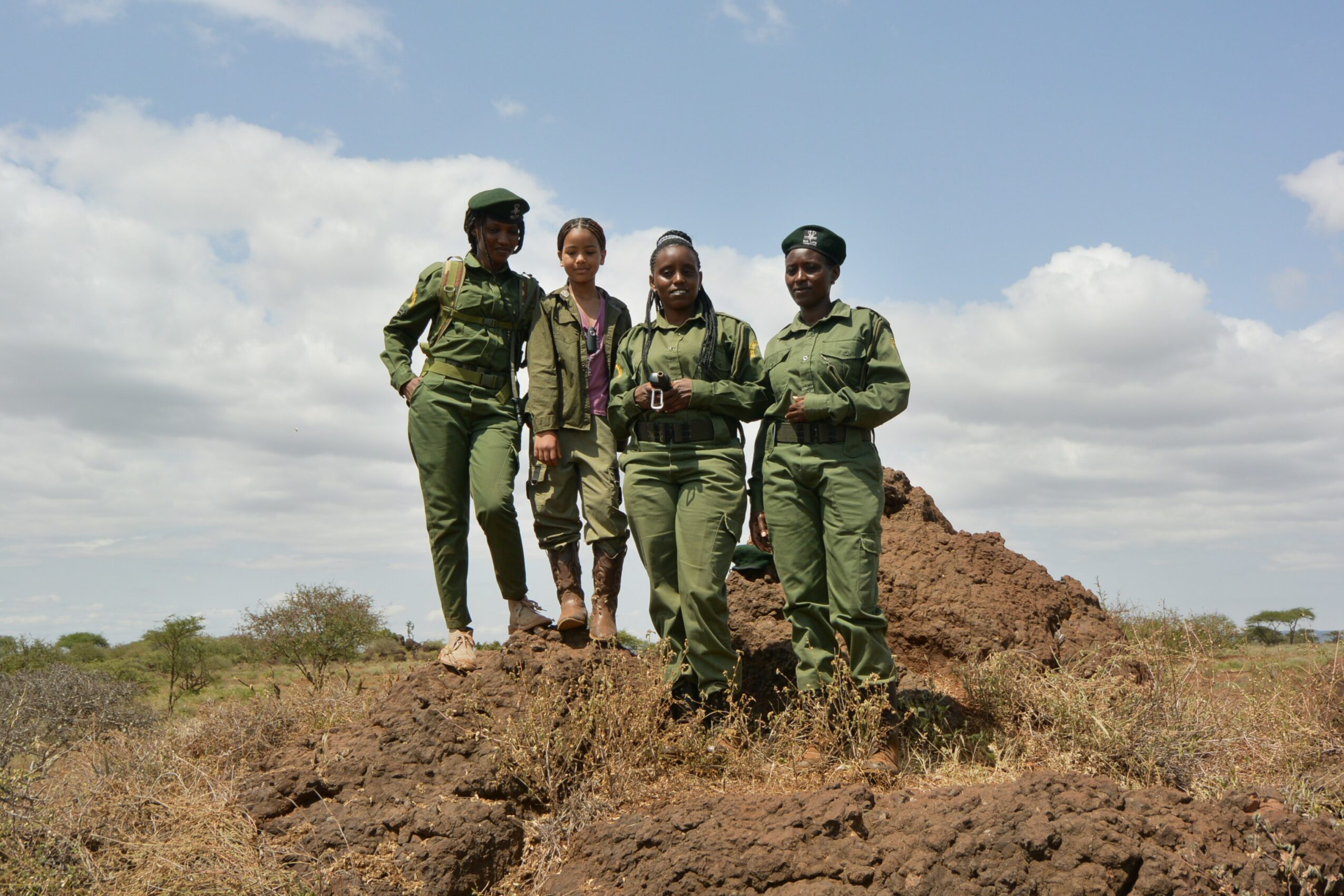 Four women in green uniforms standing on a mound of earth outdoors under a partly cloudy sky.