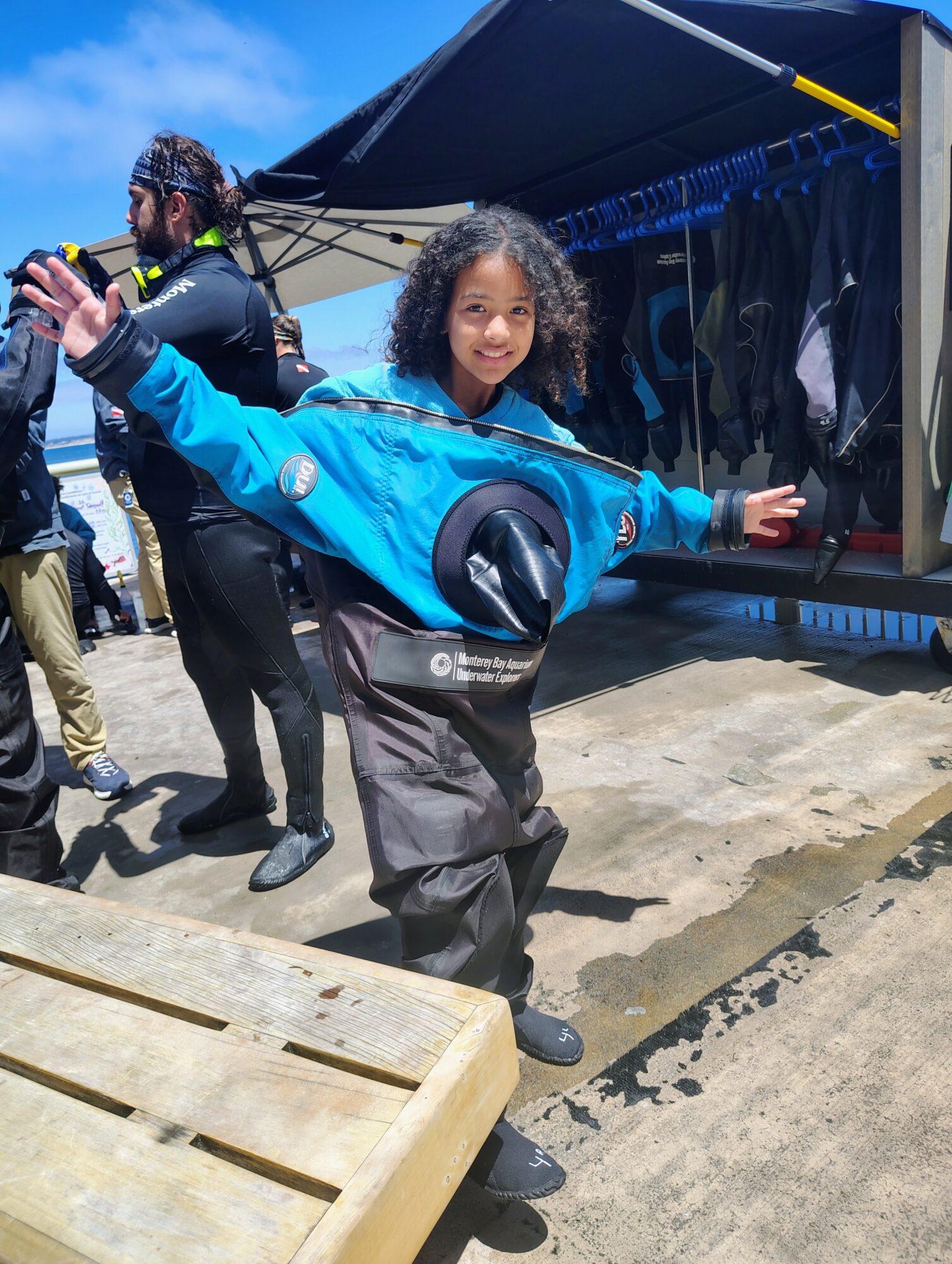 Young girl in outdoor gear smiling with arms outstretched, standing on a concrete surface near a tent and other people.