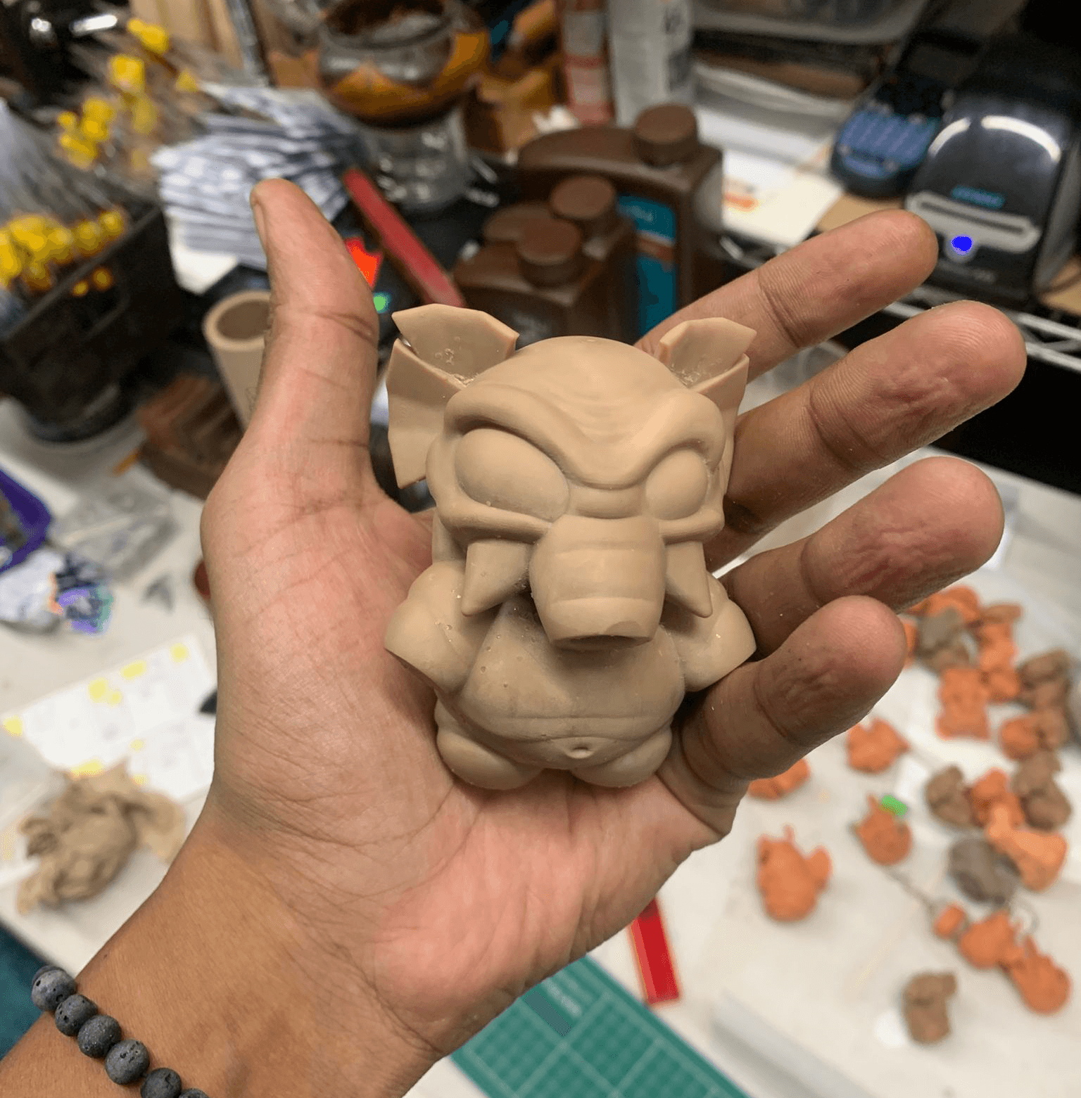 Hand holding a small, unglazed clay sculpture of a creature with large ears and a snout, surrounded by art supplies.