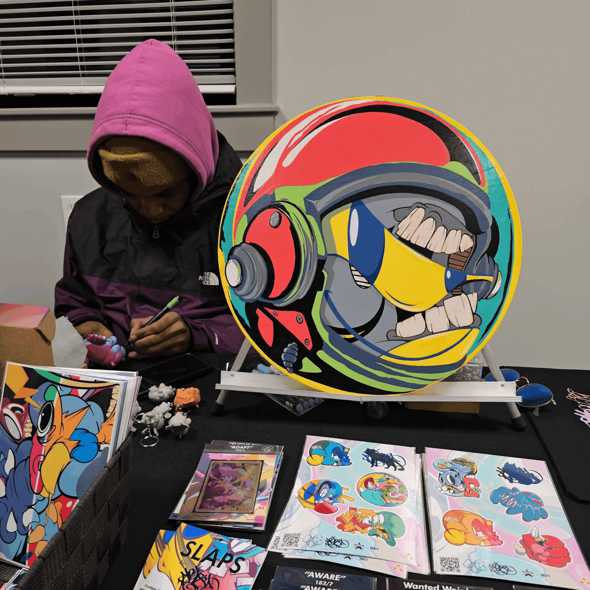 Person in pink hoodie and yellow mask working on a colorful, round artwork with a cartoon character. Various art supplies and books on table.