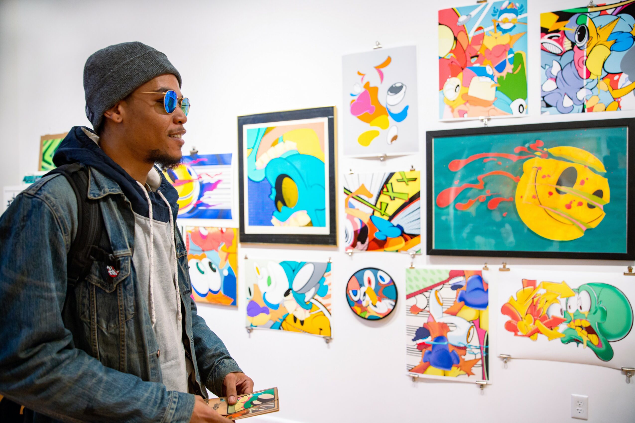 Person wearing sunglasses and a beanie looking at colorful abstract artwork on a gallery wall.