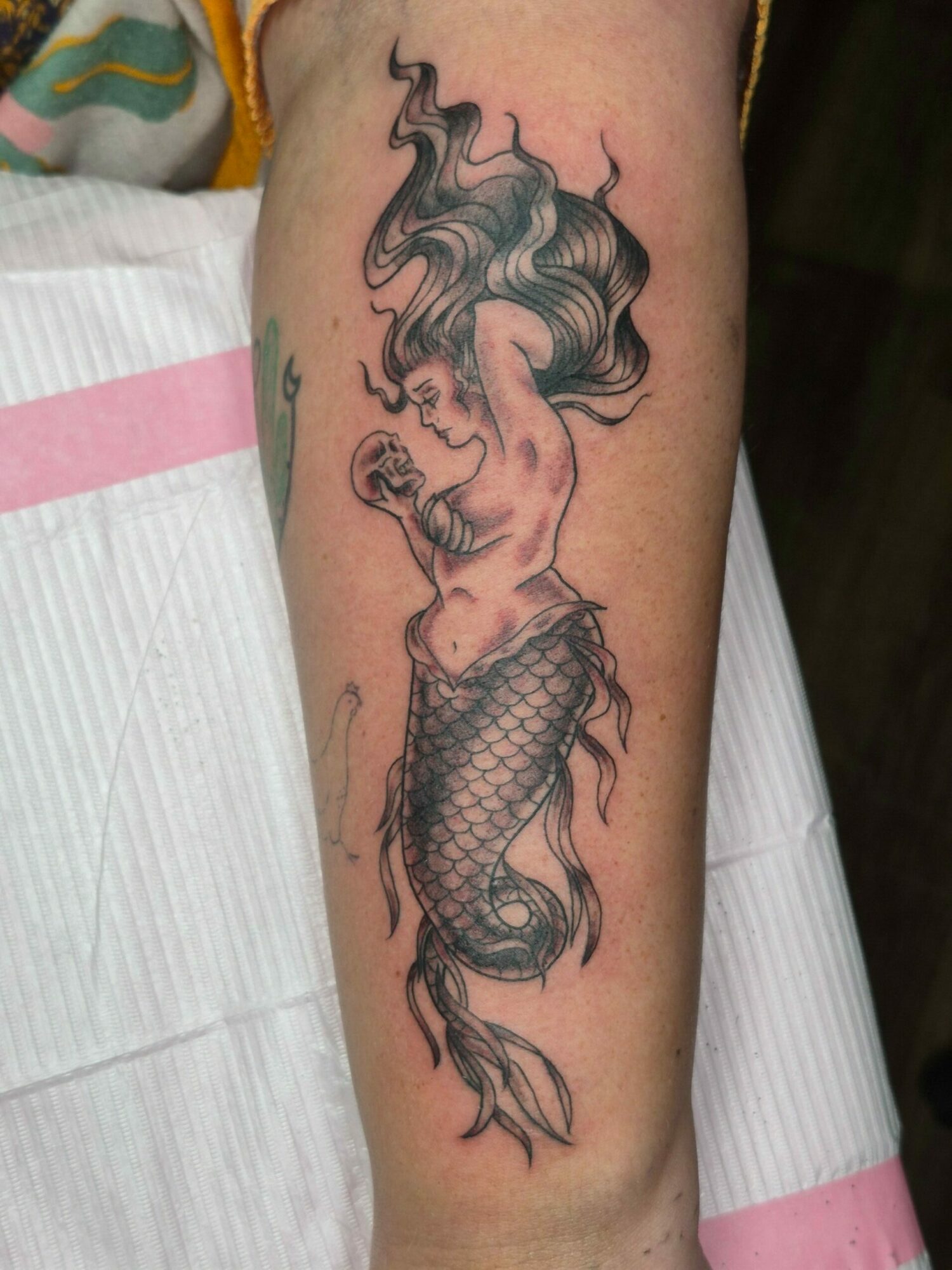 Tattoo of a mermaid with flowing hair and a fish tail on a person's arm.