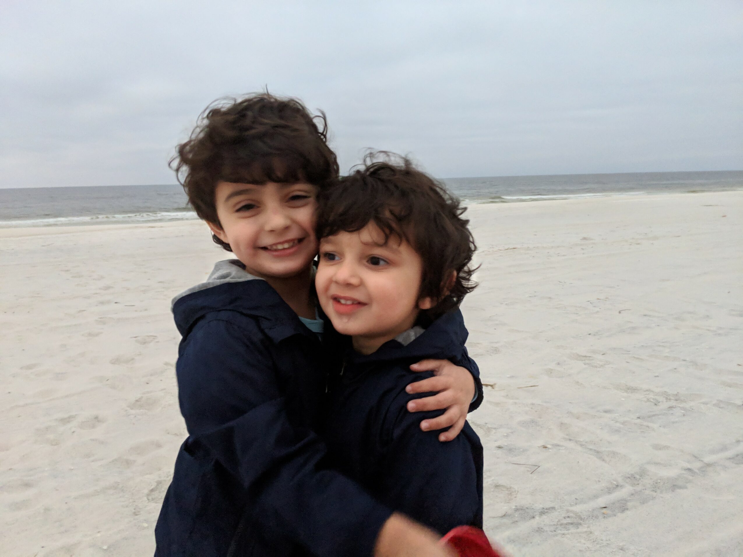 Two children hugging on a sandy beach with the ocean in the background, smiling happily.