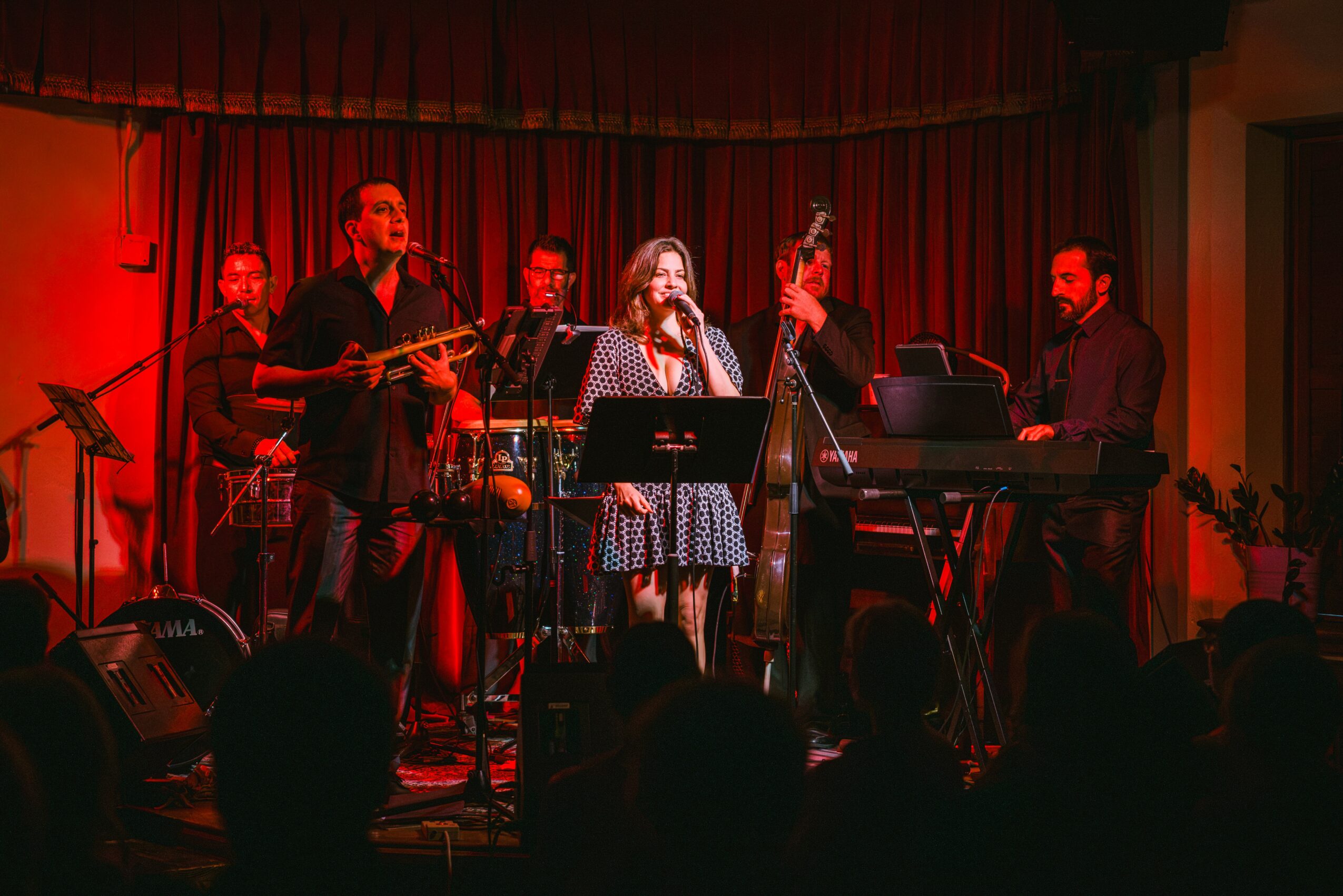 Six musicians perform on stage with red curtains, playing instruments including saxophone, keyboard, and vocals, in a dimly lit setting.