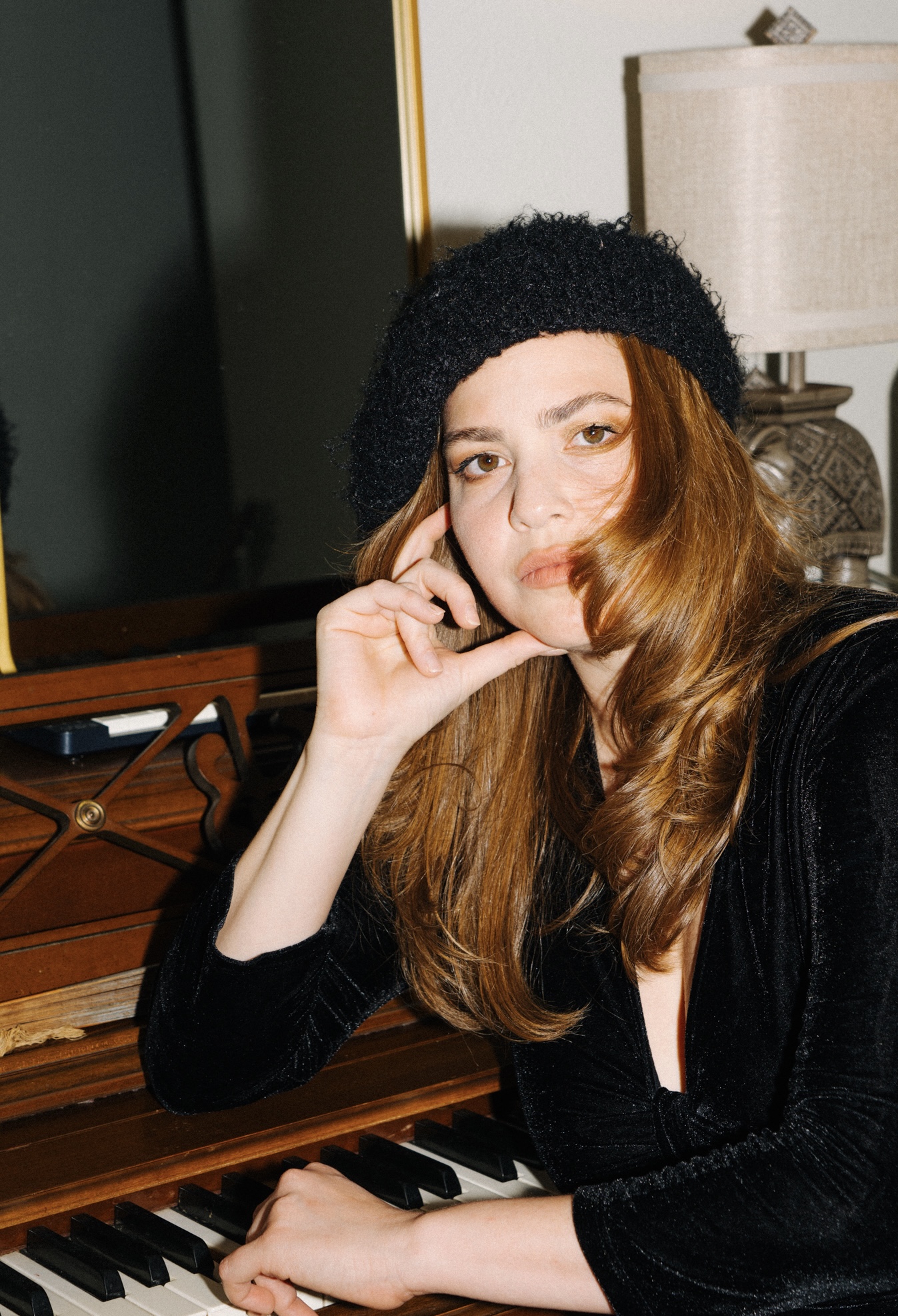 Young woman with long hair wearing a black hat and velvet top, resting her chin on her hand, sitting at a piano.